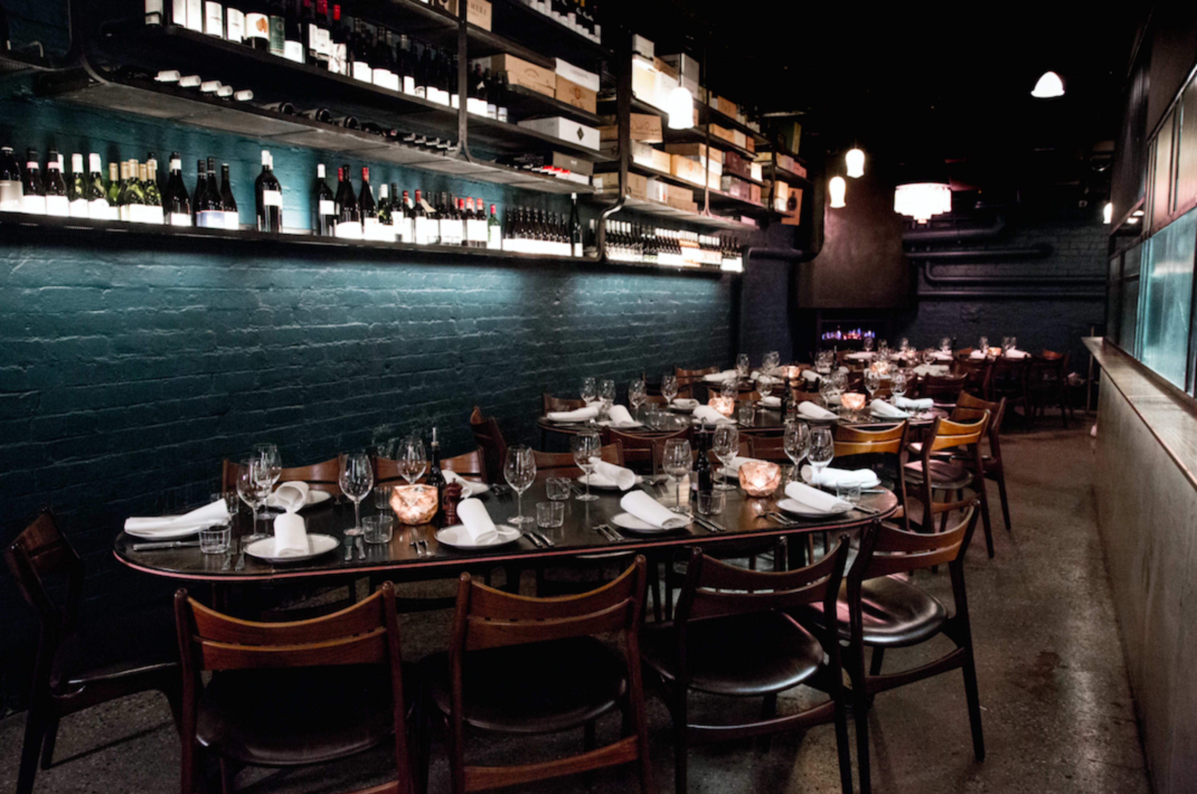 The image shows an elegantly arranged dining area in a restaurant with a long table set for multiple guests, surrounded by shelves of wine bottles.