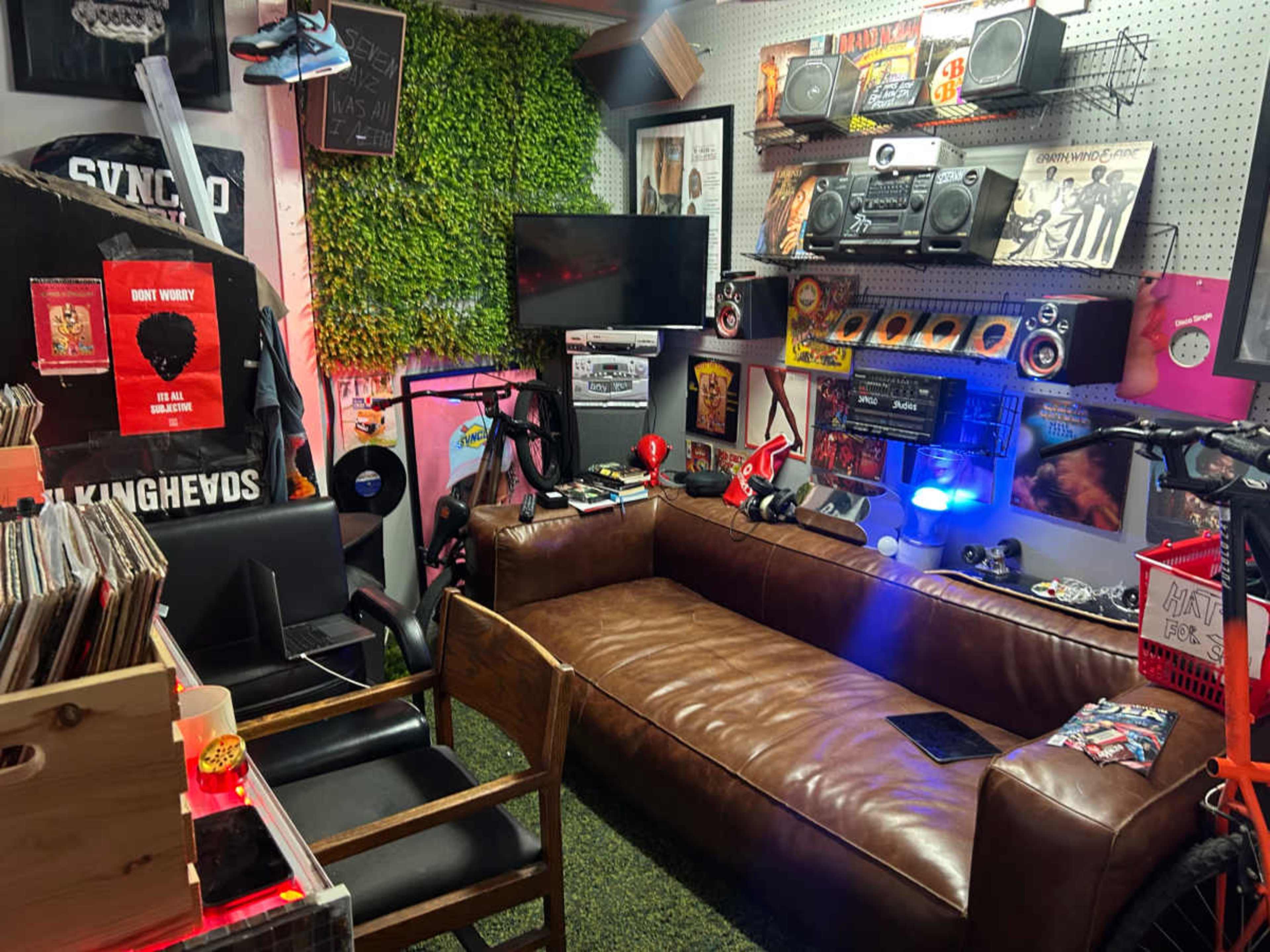 The image shows a cozy room with a leather sofa, various music equipment, vinyl records, and decorative plants on the walls.