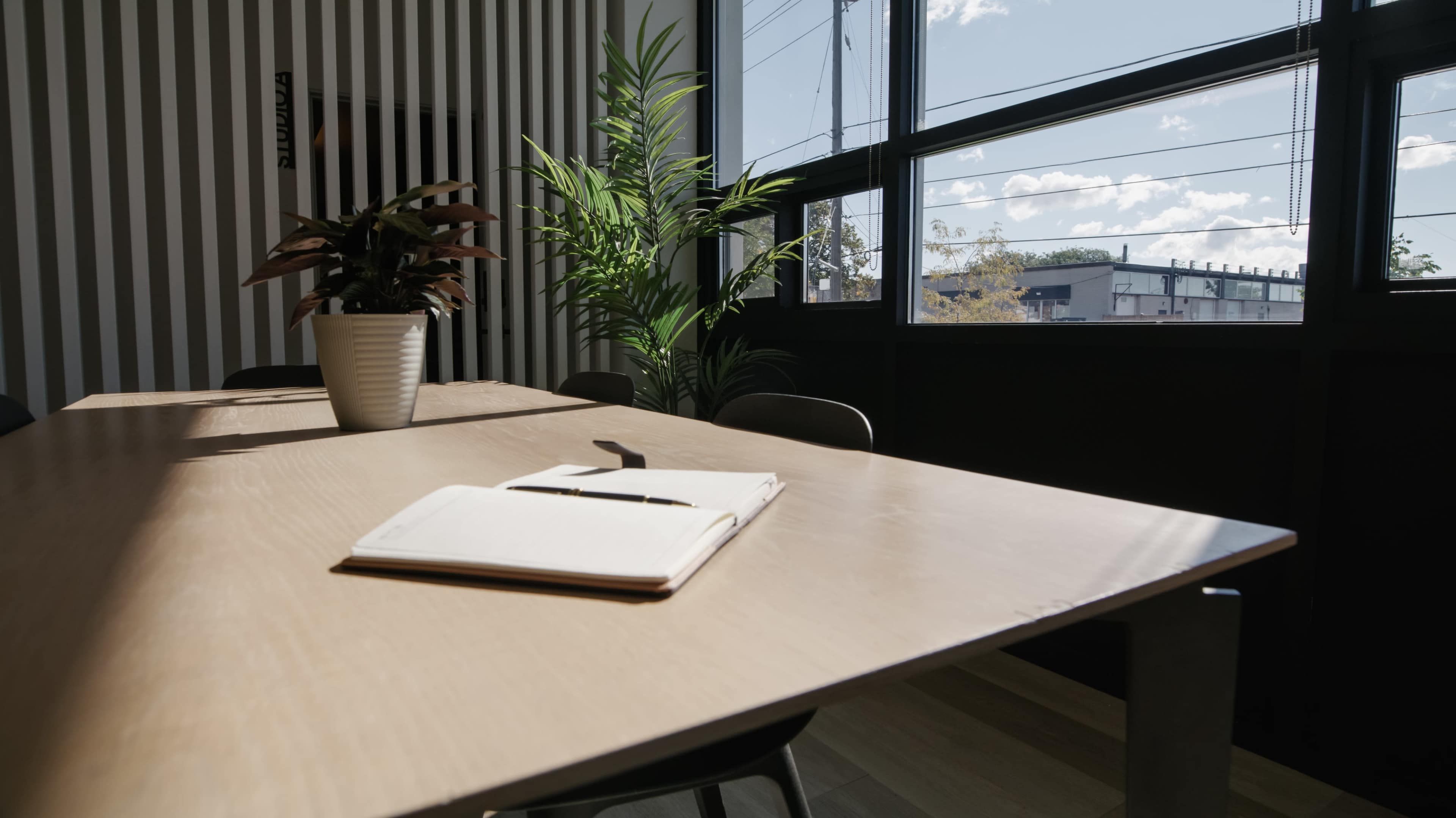 A notebook and pen are placed on a wooden conference table in a sunlit room with large windows overlooking a street.