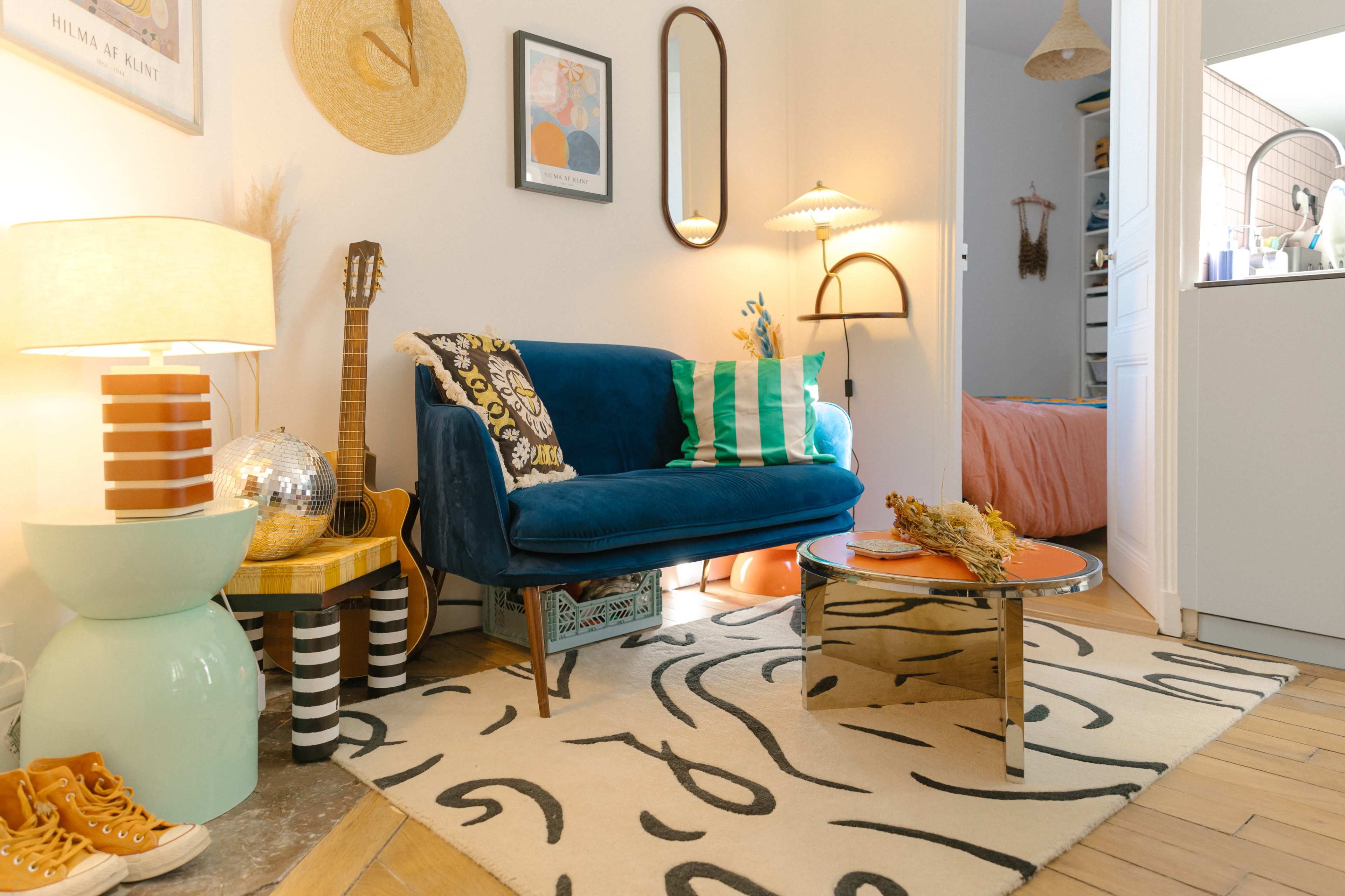 A cozy living room with a blue velvet sofa, a decorative rug, and various accessories, including a guitar, a lamp, and a round table.