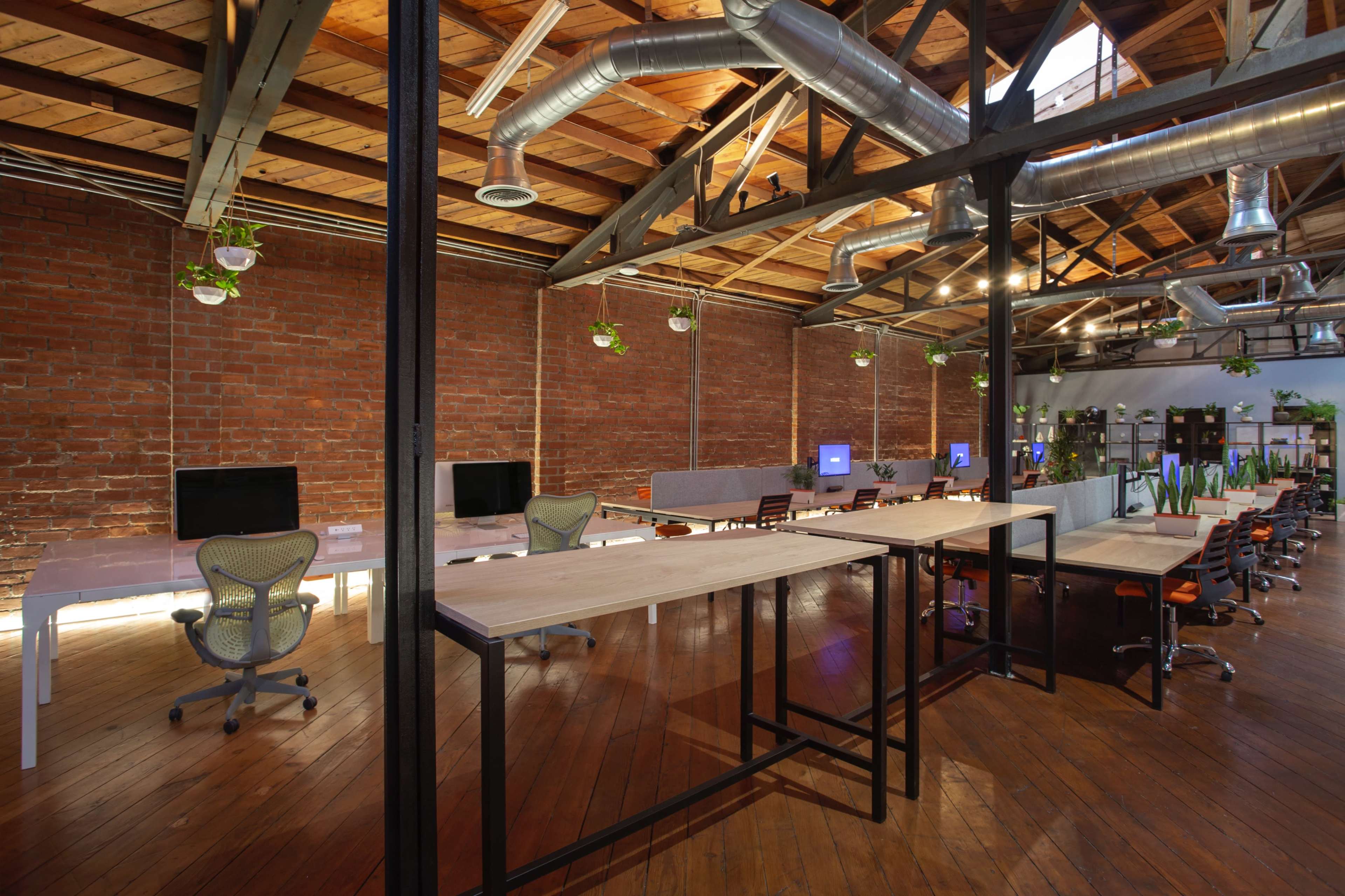 The image shows a modern open office space featuring wooden floors, exposed brick walls, and multiple workstations equipped with monitors and ergonomic chairs under a high ceiling with visible beams.