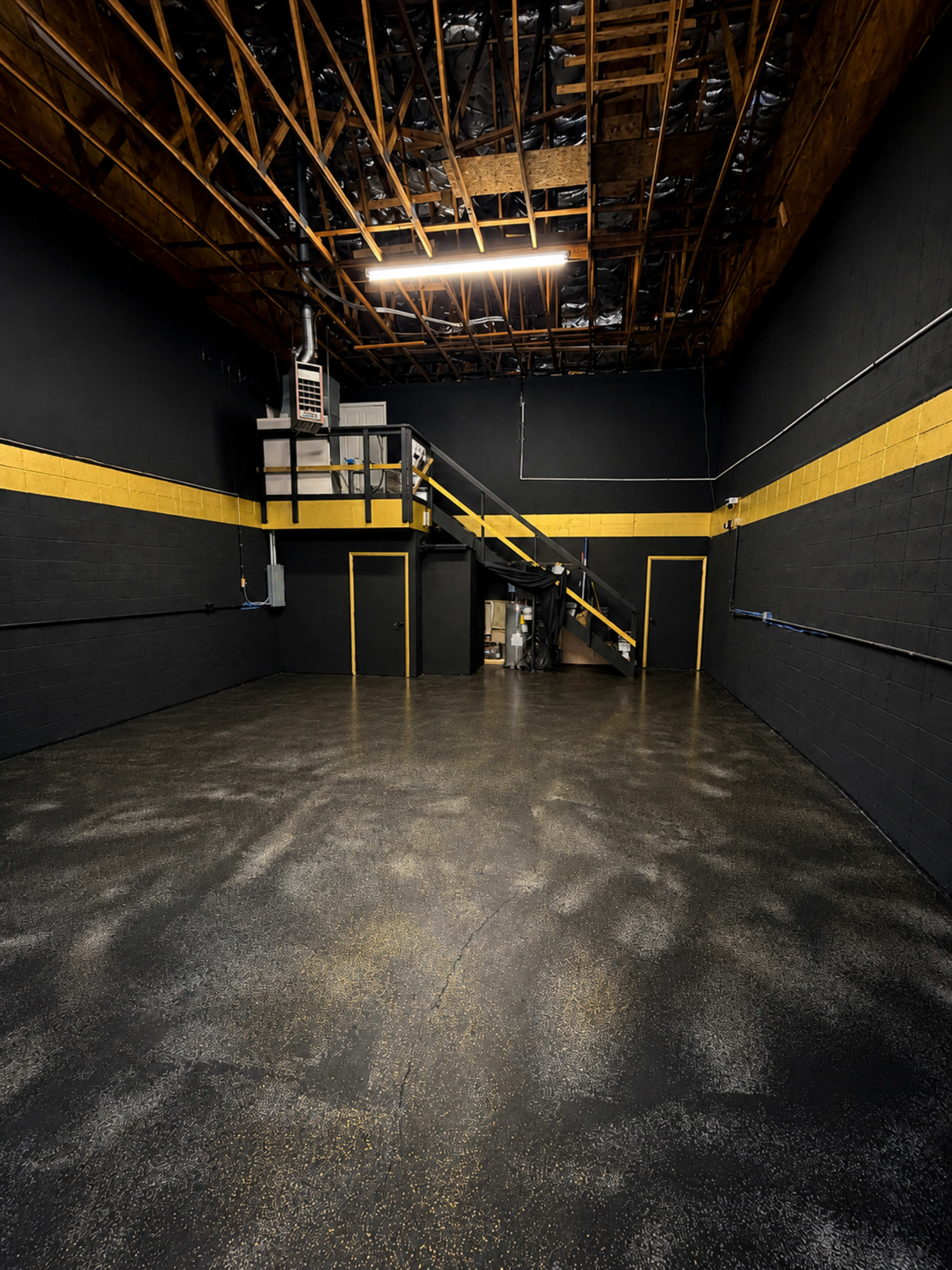 The image shows an empty industrial space with black walls, yellow stripes, a polished floor, and a staircase leading to an elevated area.