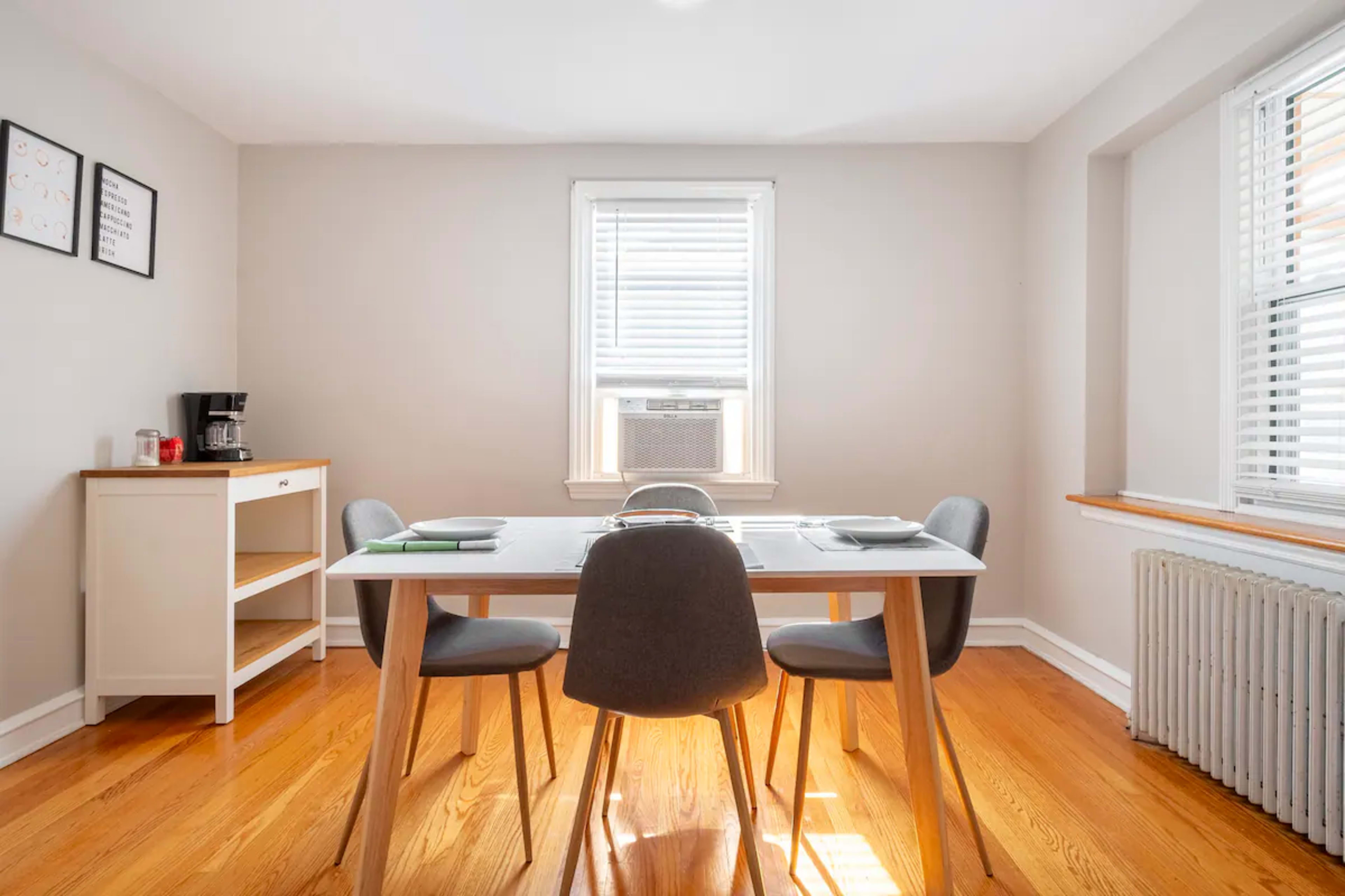 A dining area features a white table with four gray chairs, a small cabinet, and a window with an air conditioning unit.