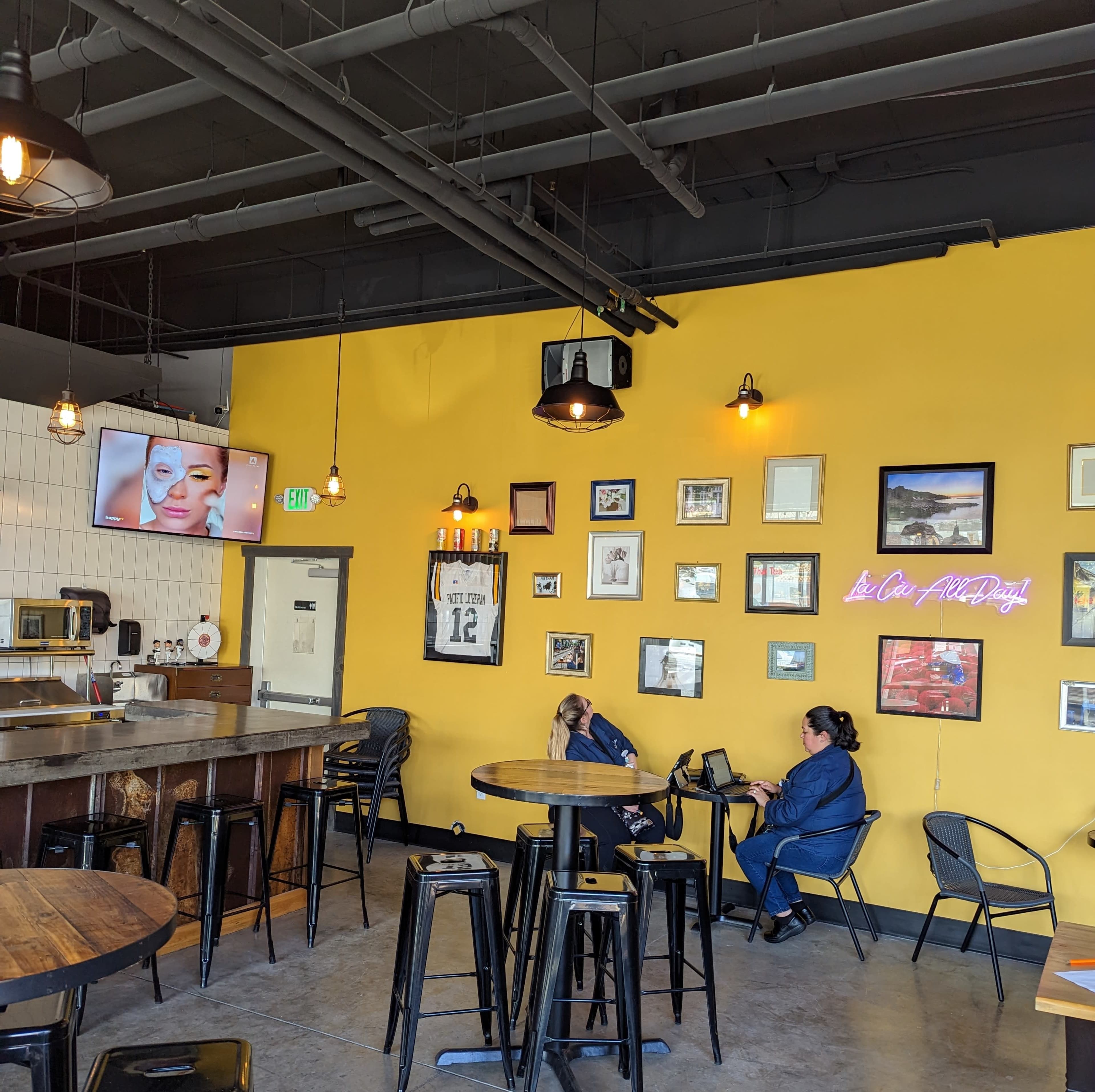 A brightly colored cafe interior with a yellow wall, a bar area with wooden tables, and two women seated at different tables.
