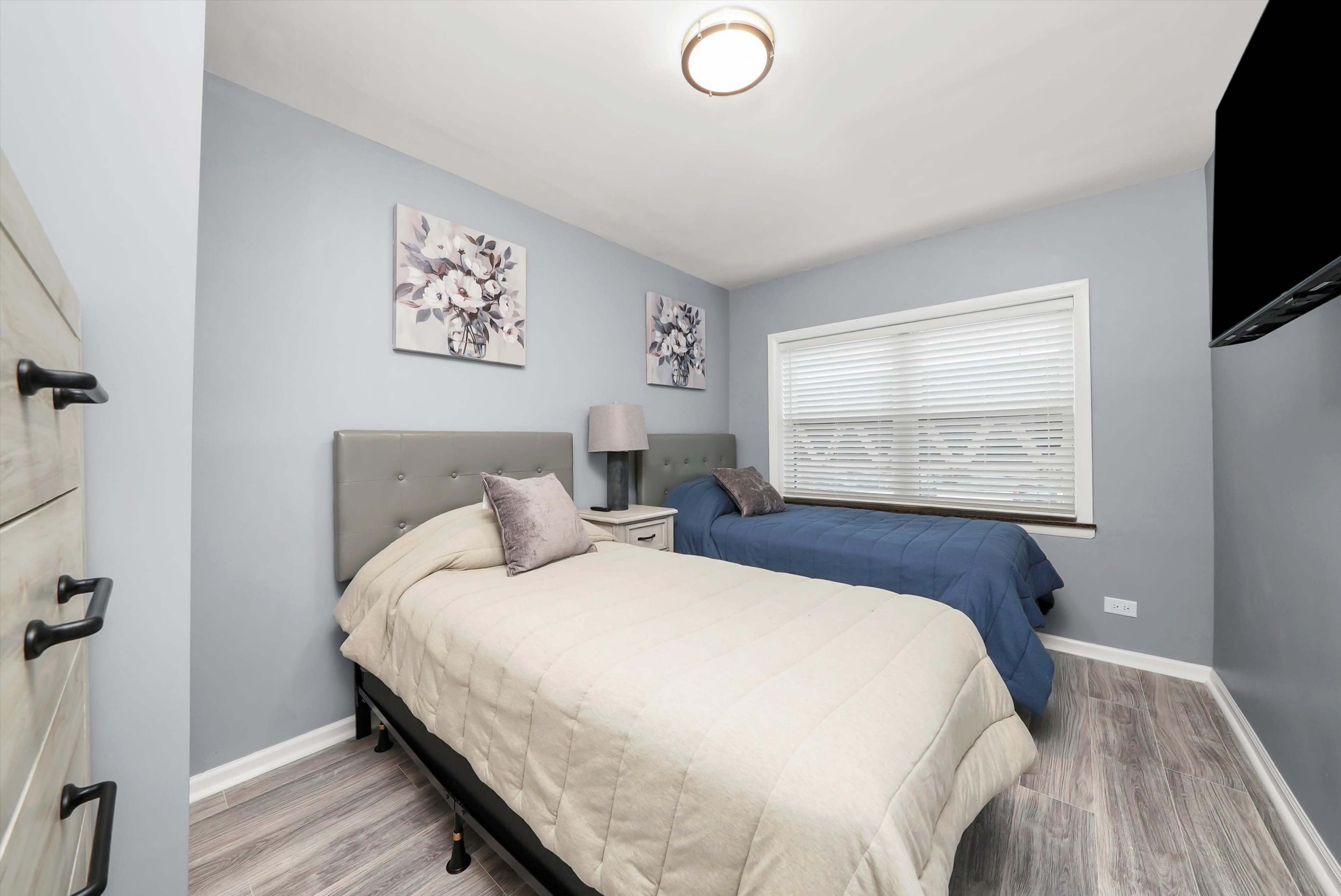 The image shows a neatly arranged bedroom featuring two twin beds, a window with blinds, wall art, and light-colored walls.