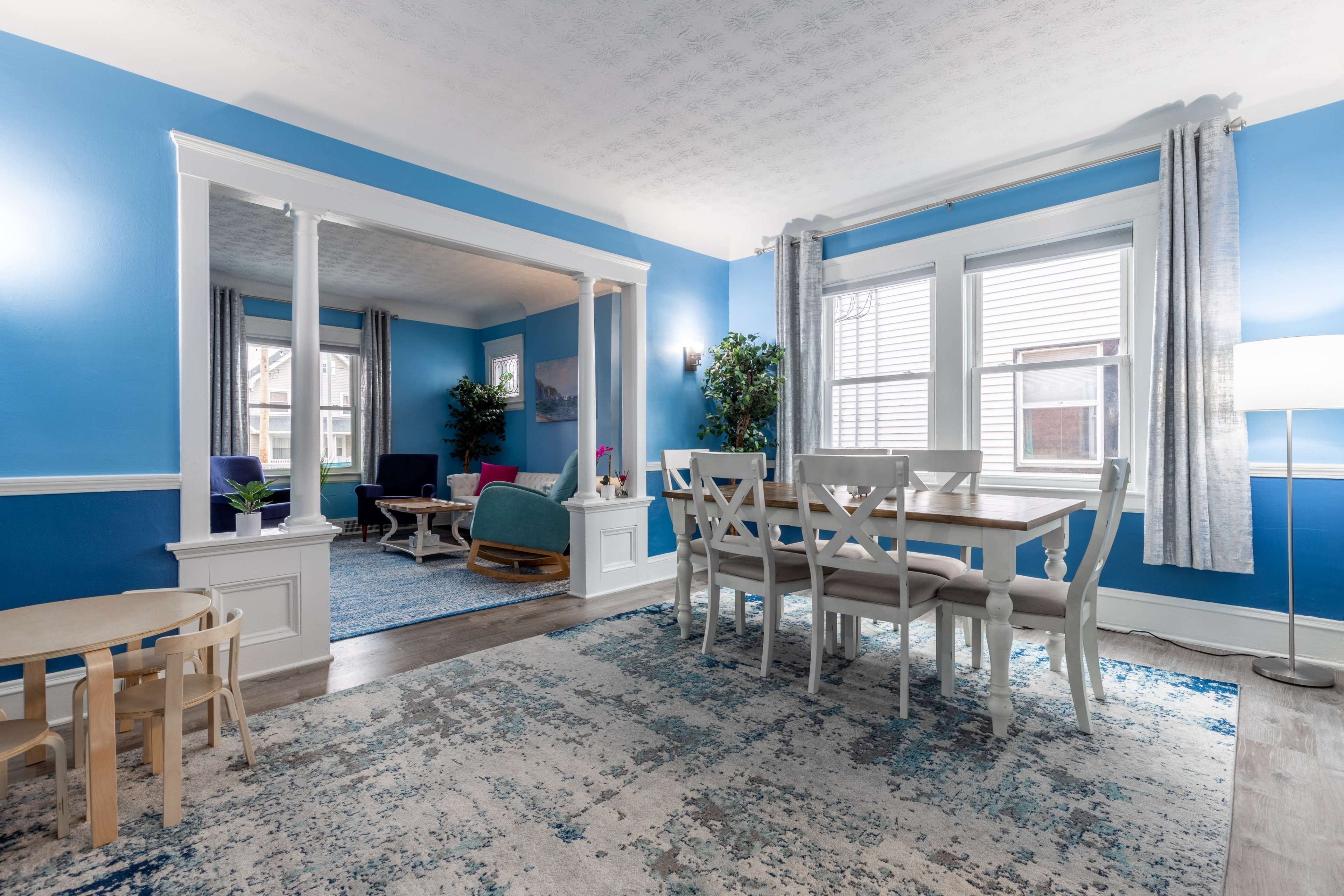 A bright living and dining area with blue walls, a wooden dining table, and seating arrangements in a separate sitting space.