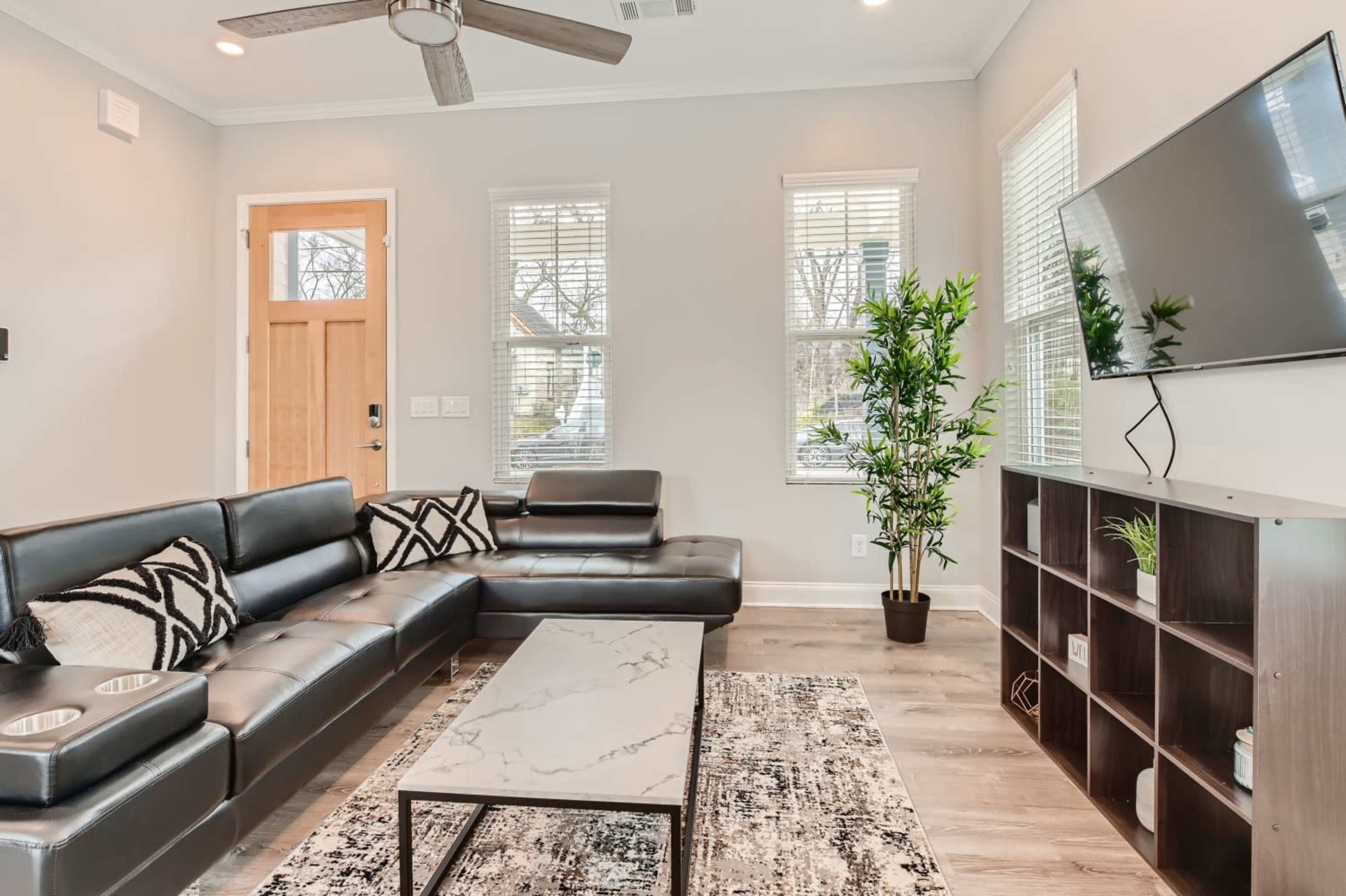 A modern living room with a black sectional sofa, a marble coffee table, a television mounted on the wall, and large windows allowing natural light.
