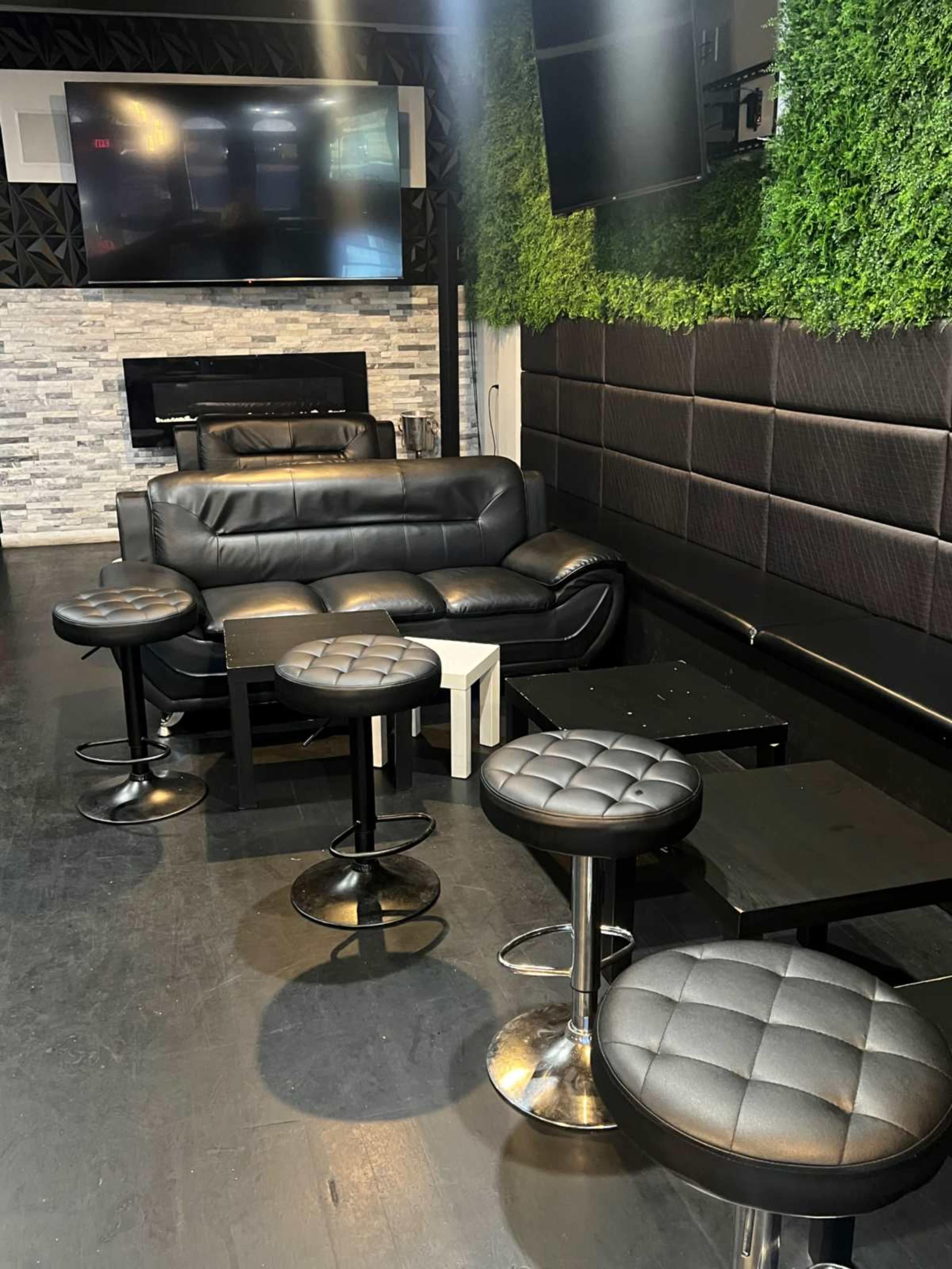 The image shows a modern lounge area with a black leather sofa, several high-top stools, and a wall covered in green foliage.