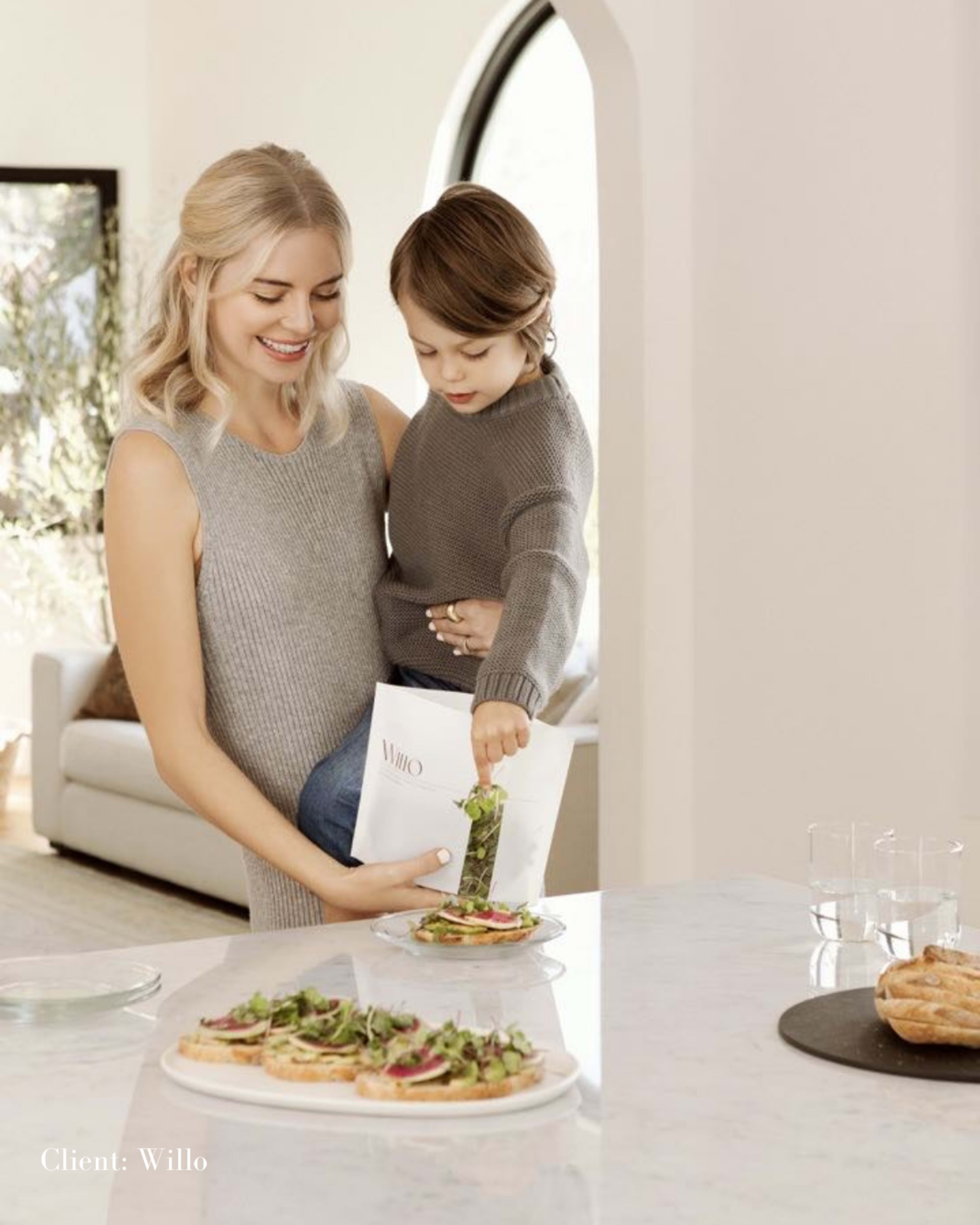 A woman holds a container while a young boy reaches inside to grab some greens on a marble kitchen counter with food plates arranged nearby.
