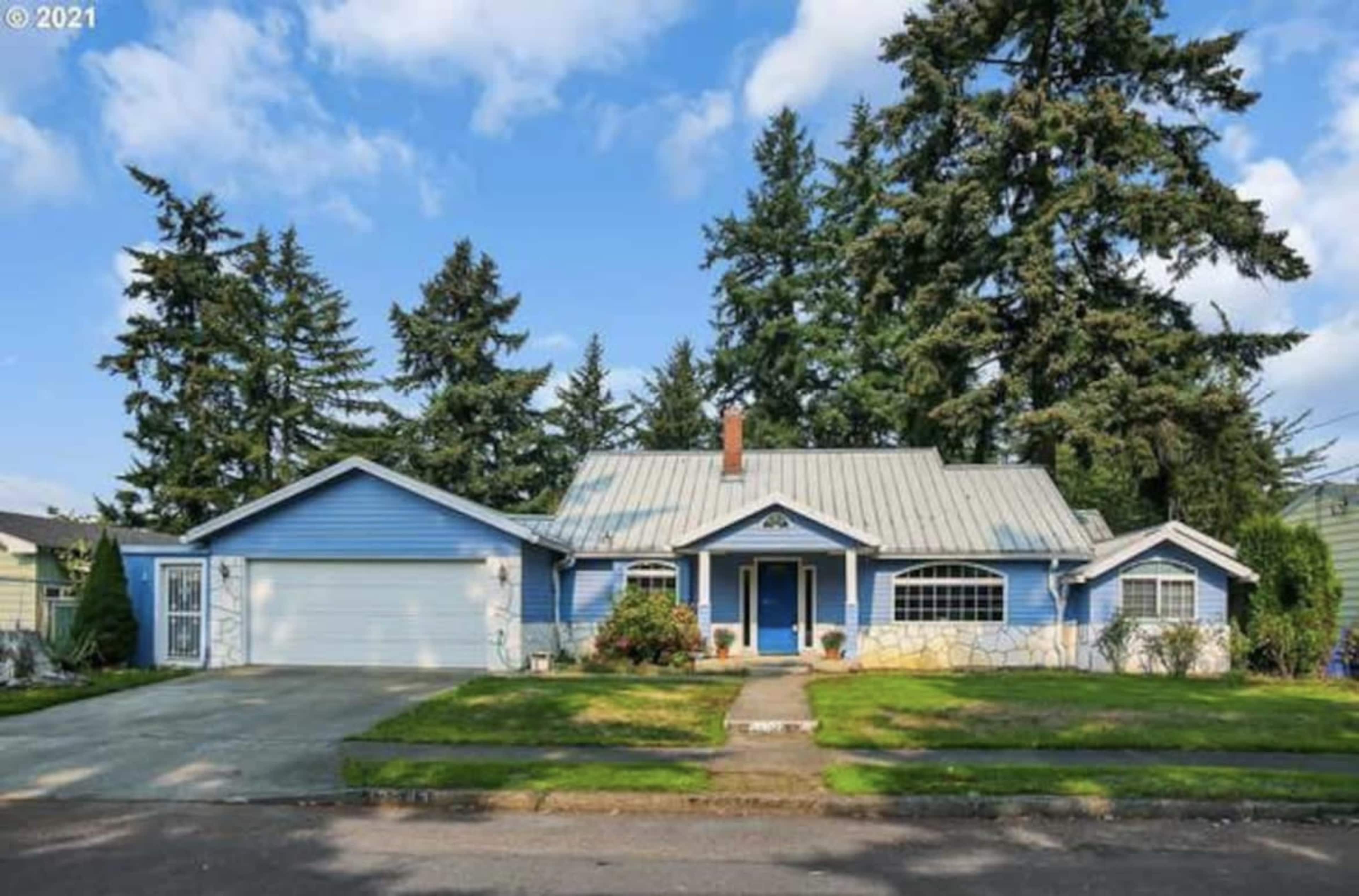 A single-story blue house with a metal roof, surrounded by well-maintained lawns and tall trees.