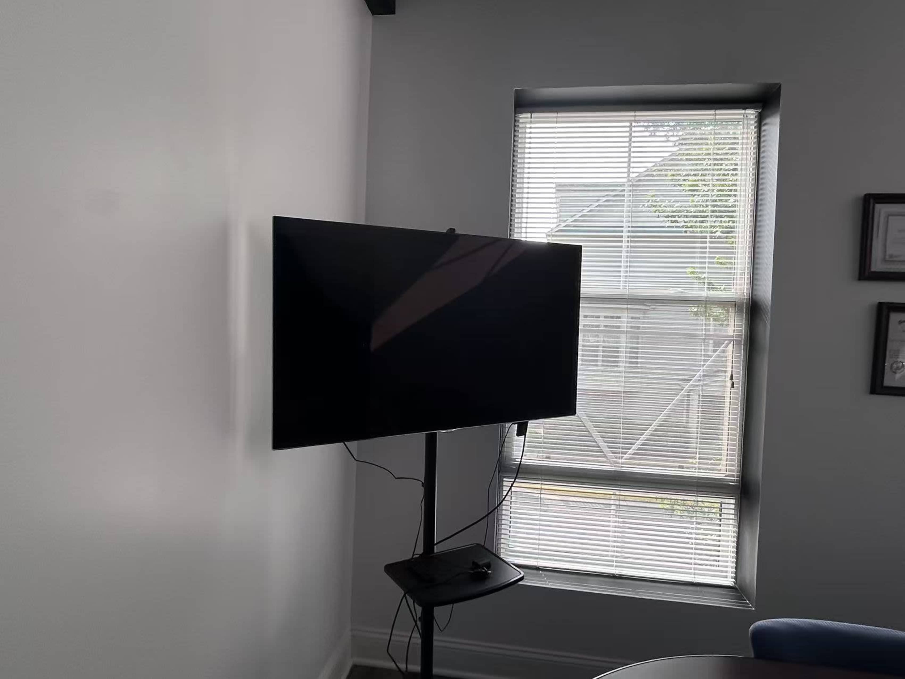 A wall-mounted television is positioned near a window with blinds in a room.