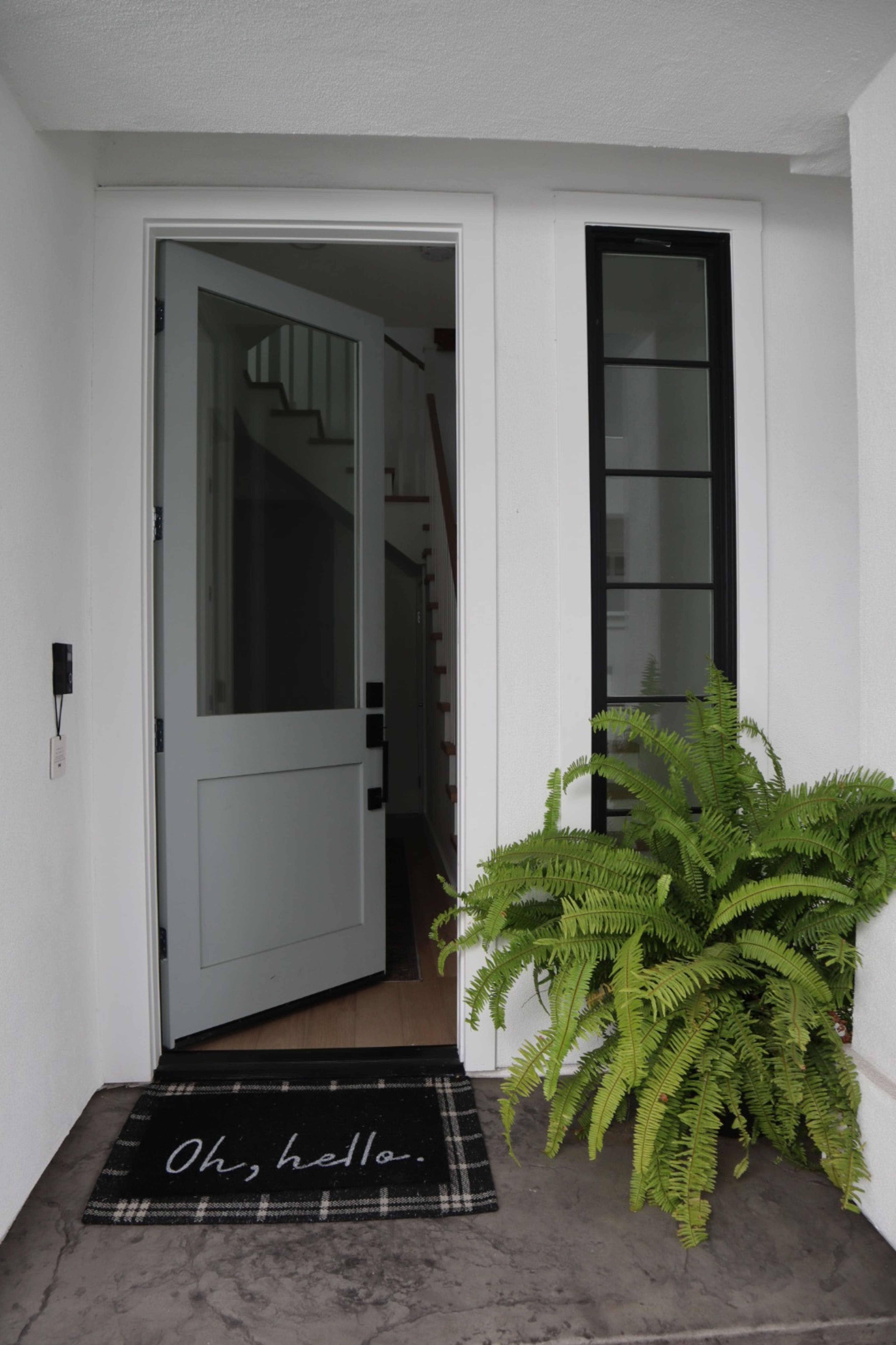 An open front door with a welcome mat that says "Oh, hello," and a potted fern beside the entrance.