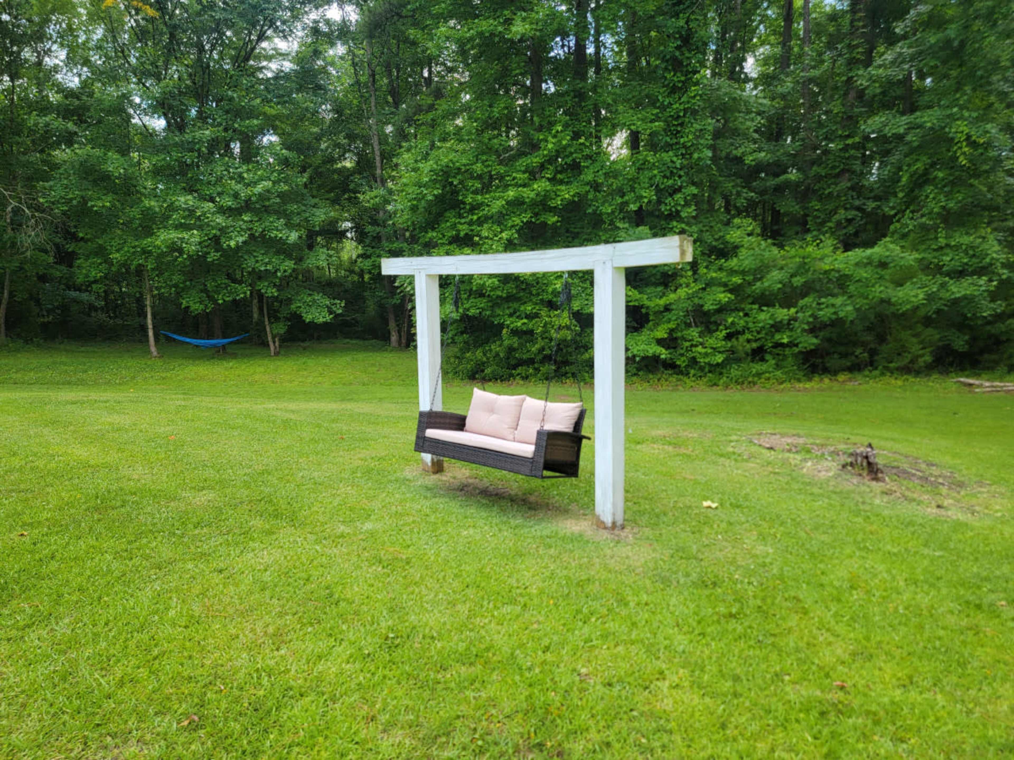 A wooden swing with a cushion hangs from a white frame in a grassy clearing surrounded by trees.