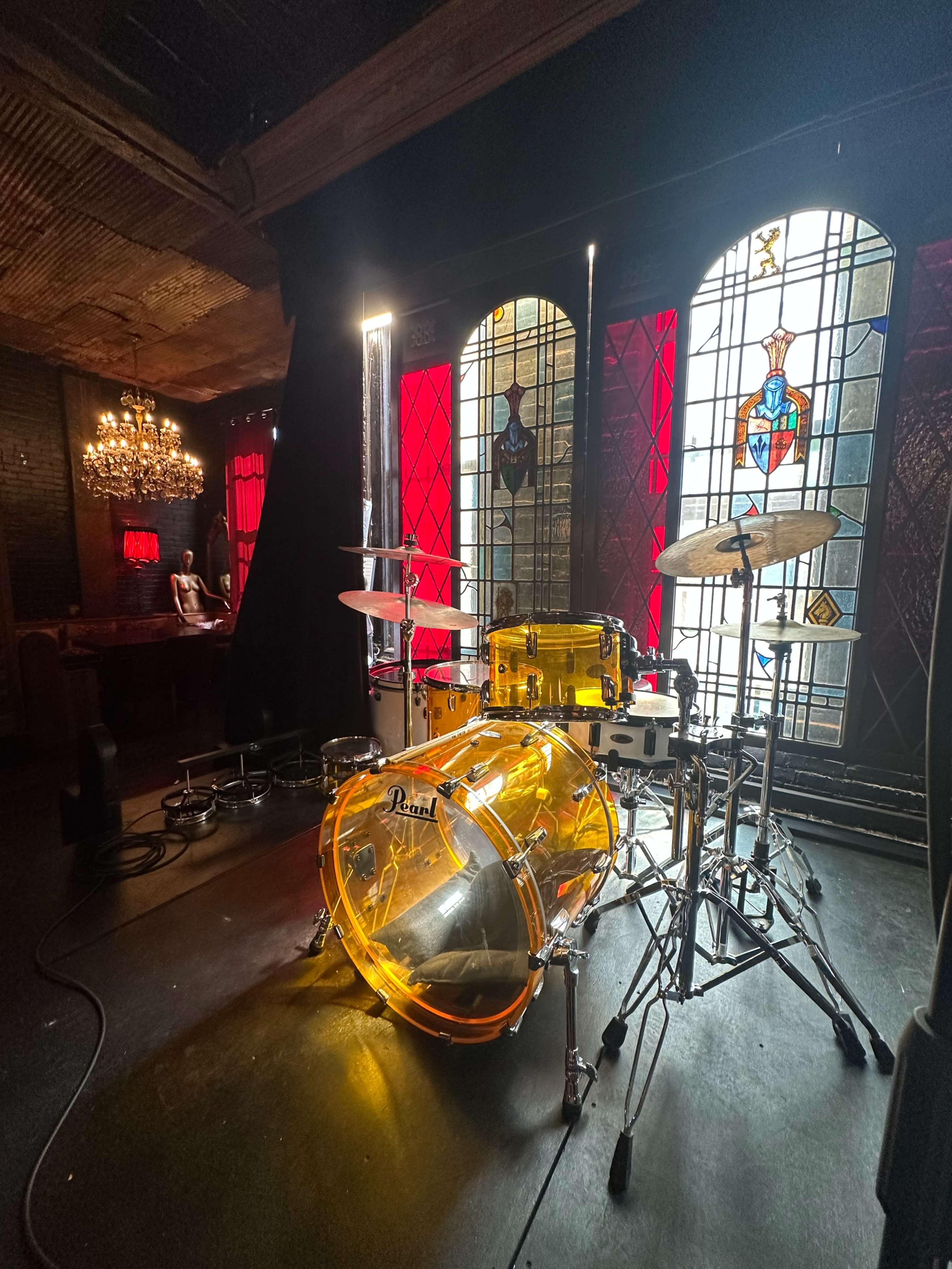 A transparent yellow drum kit is set up on a stage in front of stained glass windows with red accents.