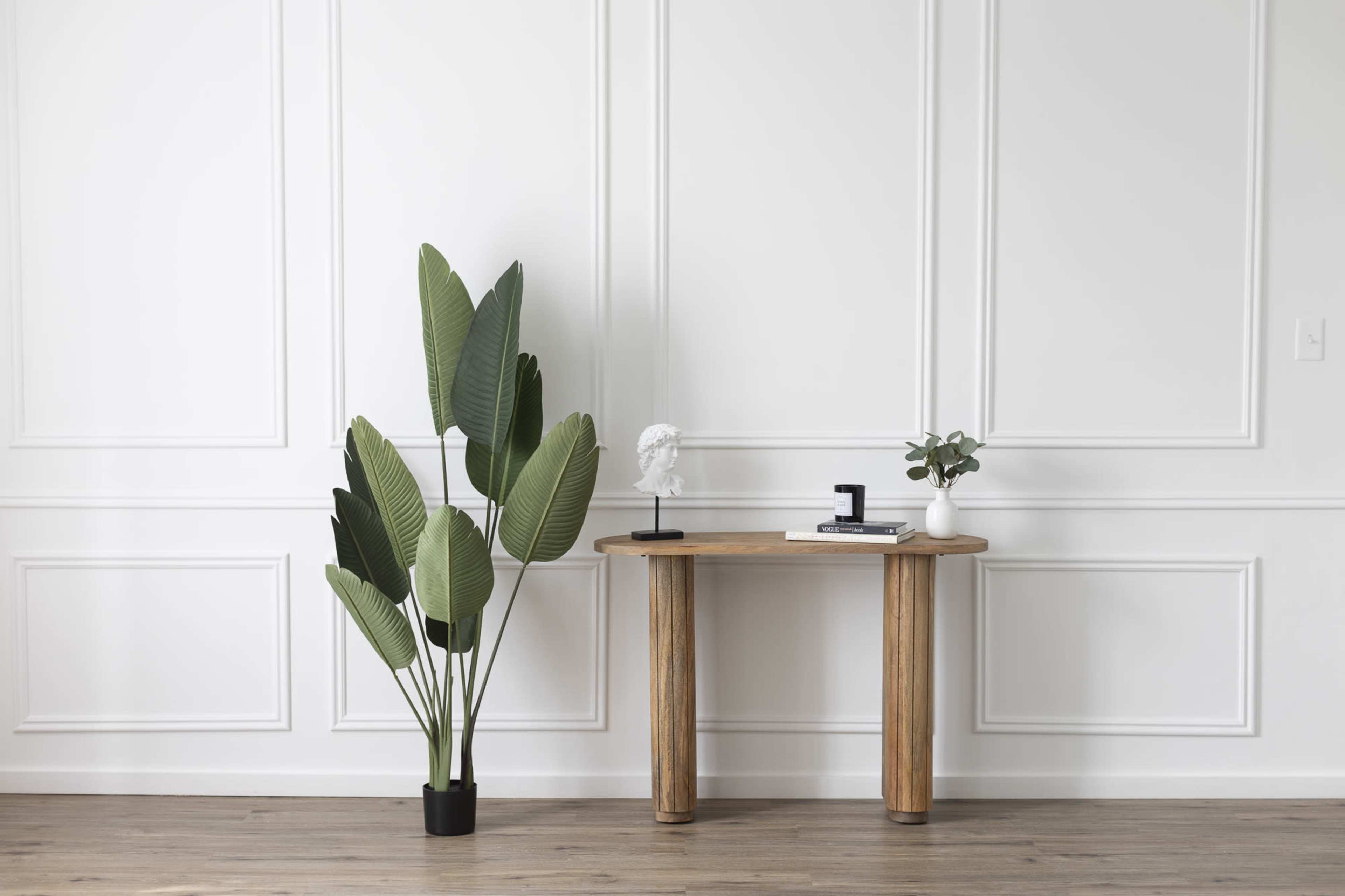 A wooden console table with a black decorative object, a small plant, and a sculpture is positioned against a white paneled wall, alongside a large artificial plant in a pot.