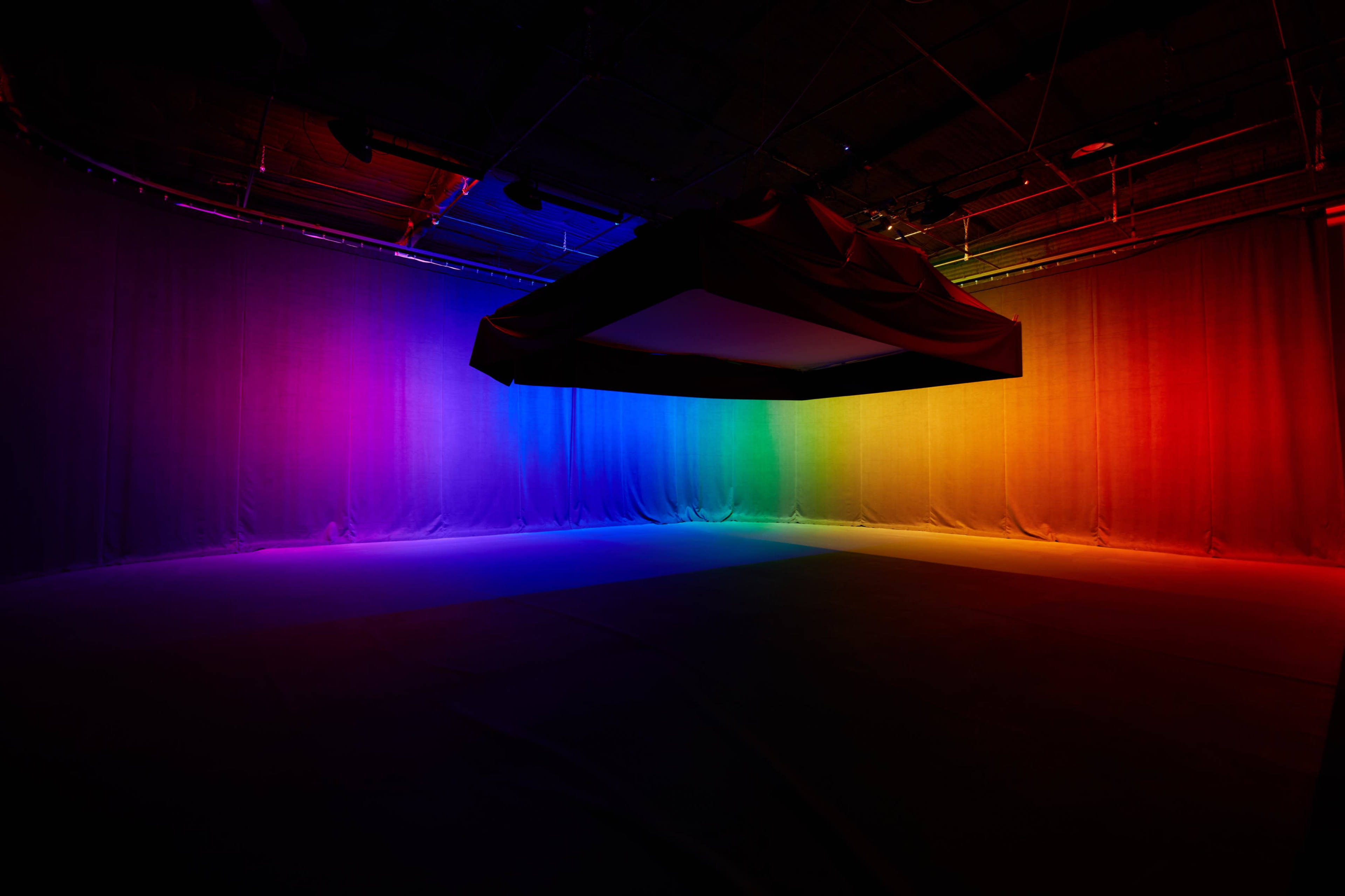 A dark room features a large, suspended rectangular object with colorful light gradients illuminating the walls.