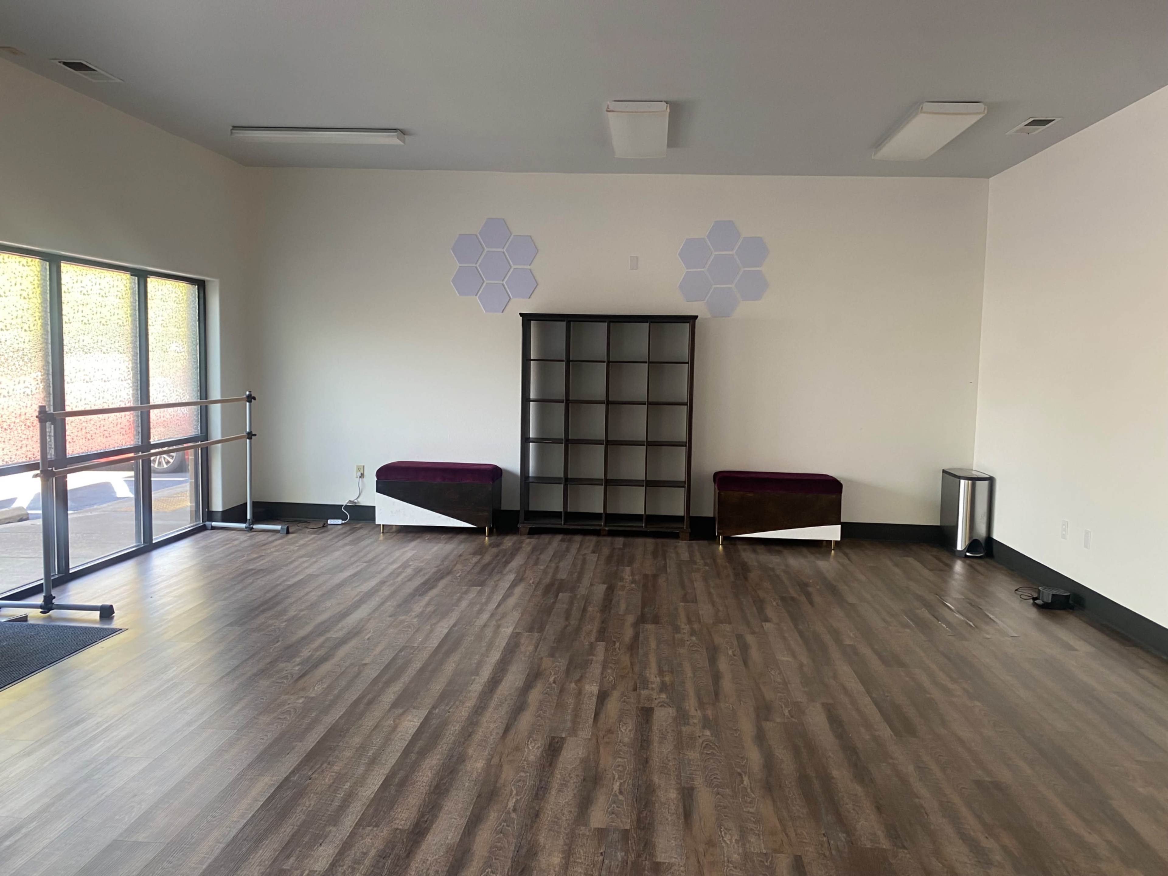 The image shows an empty room with wooden flooring, a bookshelf against one wall, two benches with purple upholstery, and a large window allowing natural light.