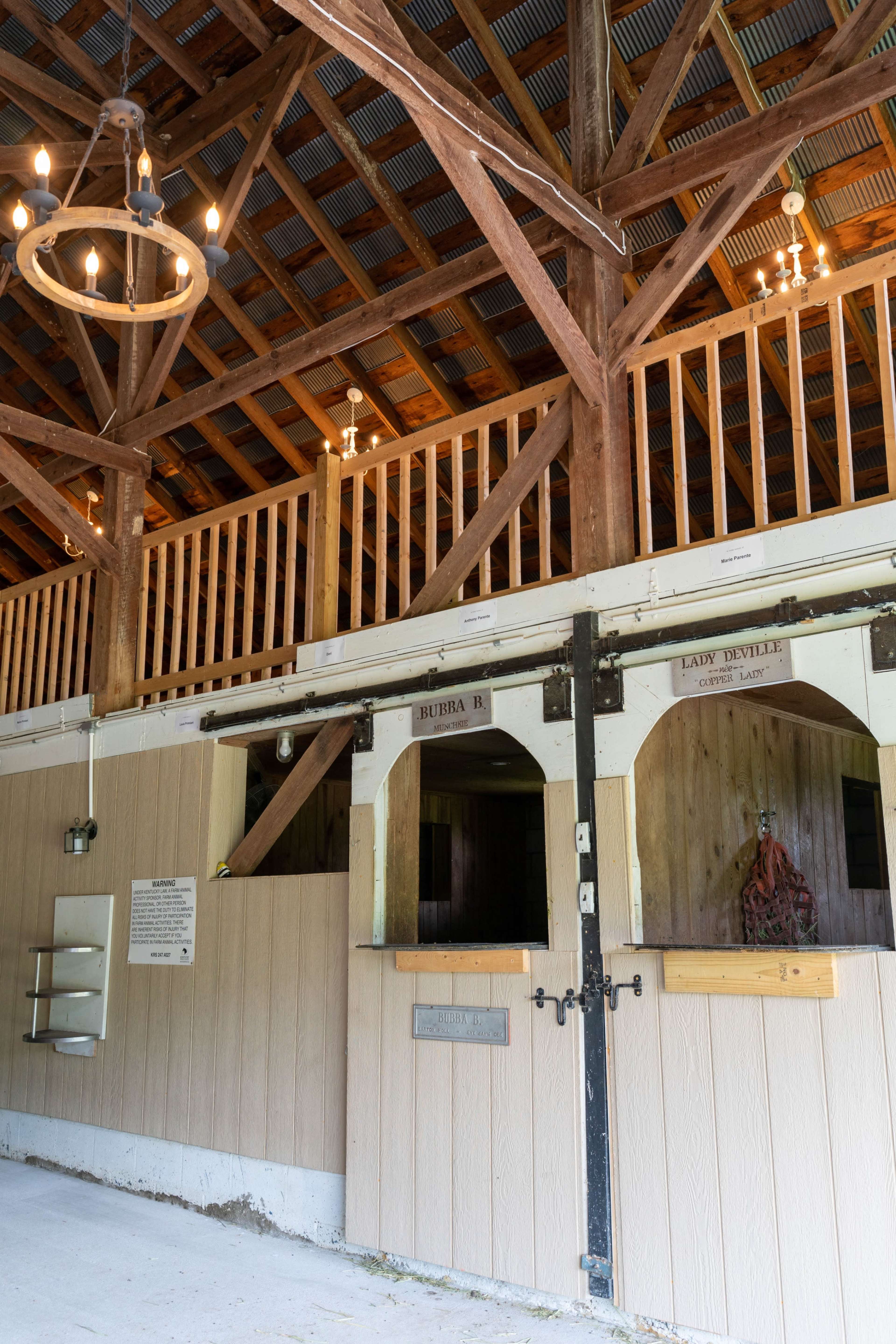 The interior of a barn featuring wooden beams, a second-story balcony, and a pair of horse stalls with names labeled above them.