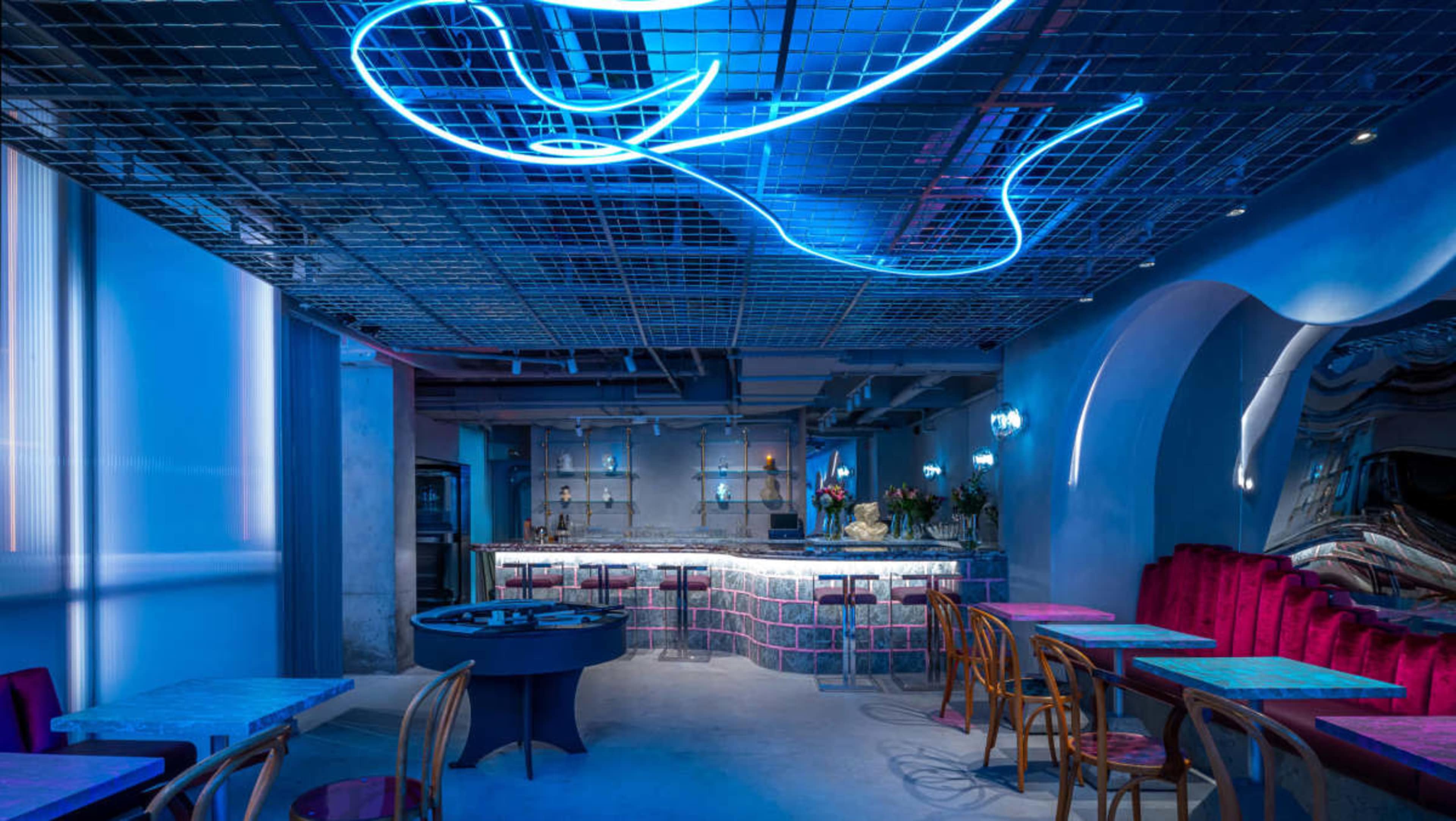 The image shows a modern restaurant interior with vibrant blue lighting, a bar at the back, and various colorful tables and chairs arranged throughout the space.