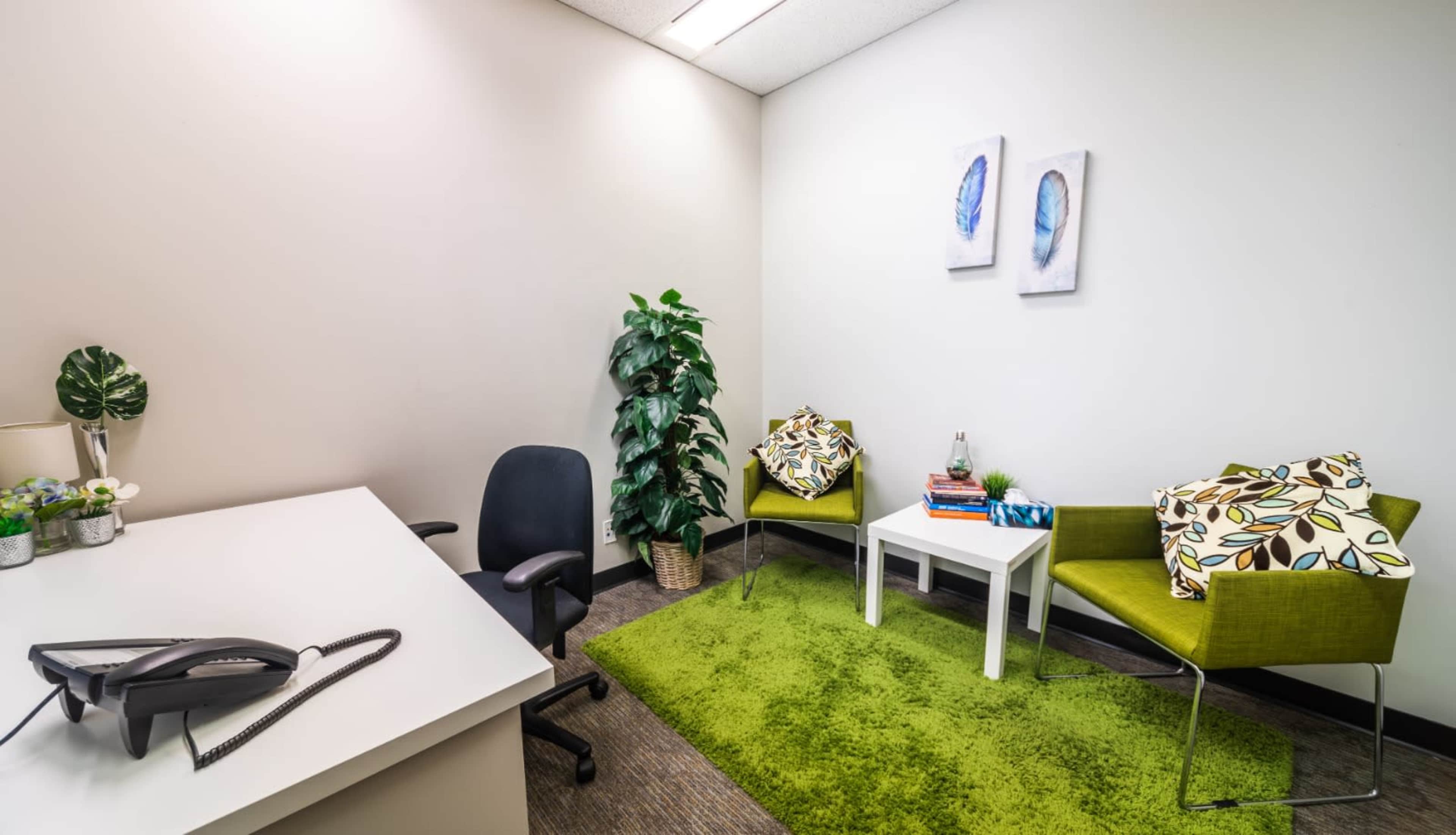 A small office space with a desk, a chair, two green accent chairs with patterned cushions, a coffee table with books, a plant, and two framed artworks on the wall.