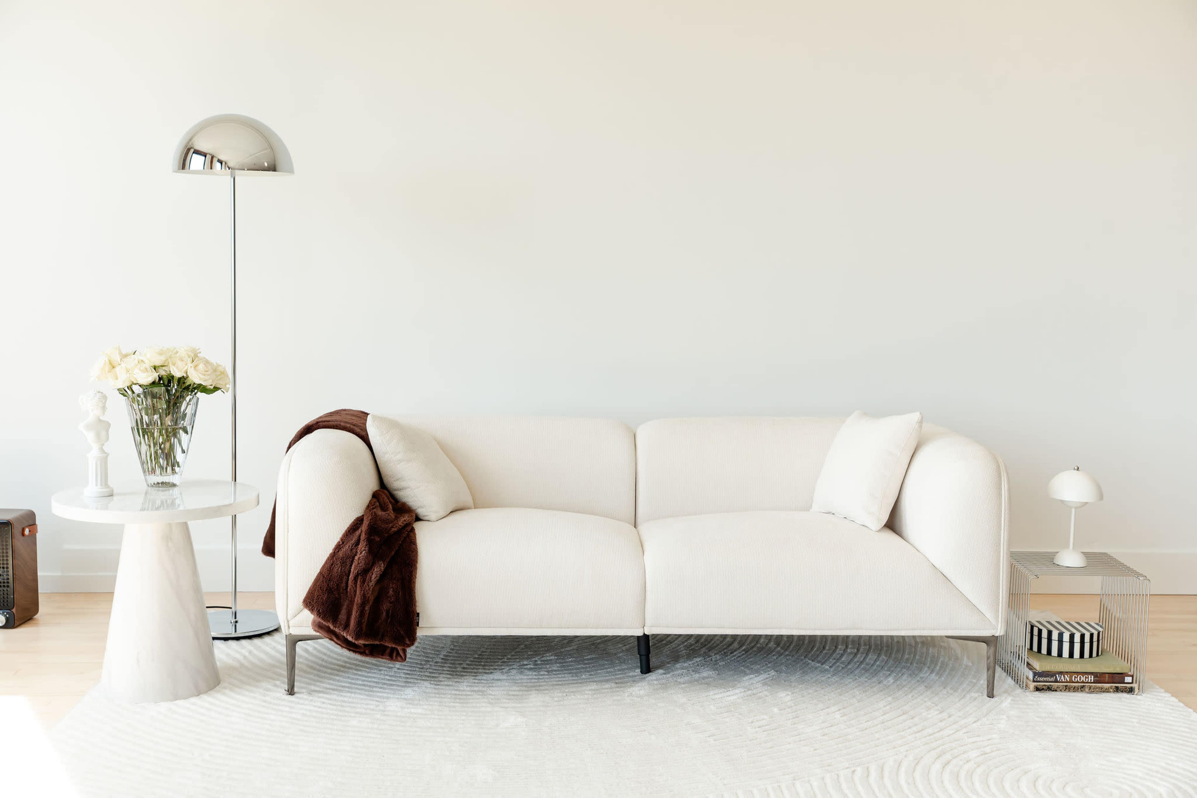 The image shows a modern, minimalistic living room featuring a light-colored sofa, a small table with flowers, a floor lamp, and a round area rug.