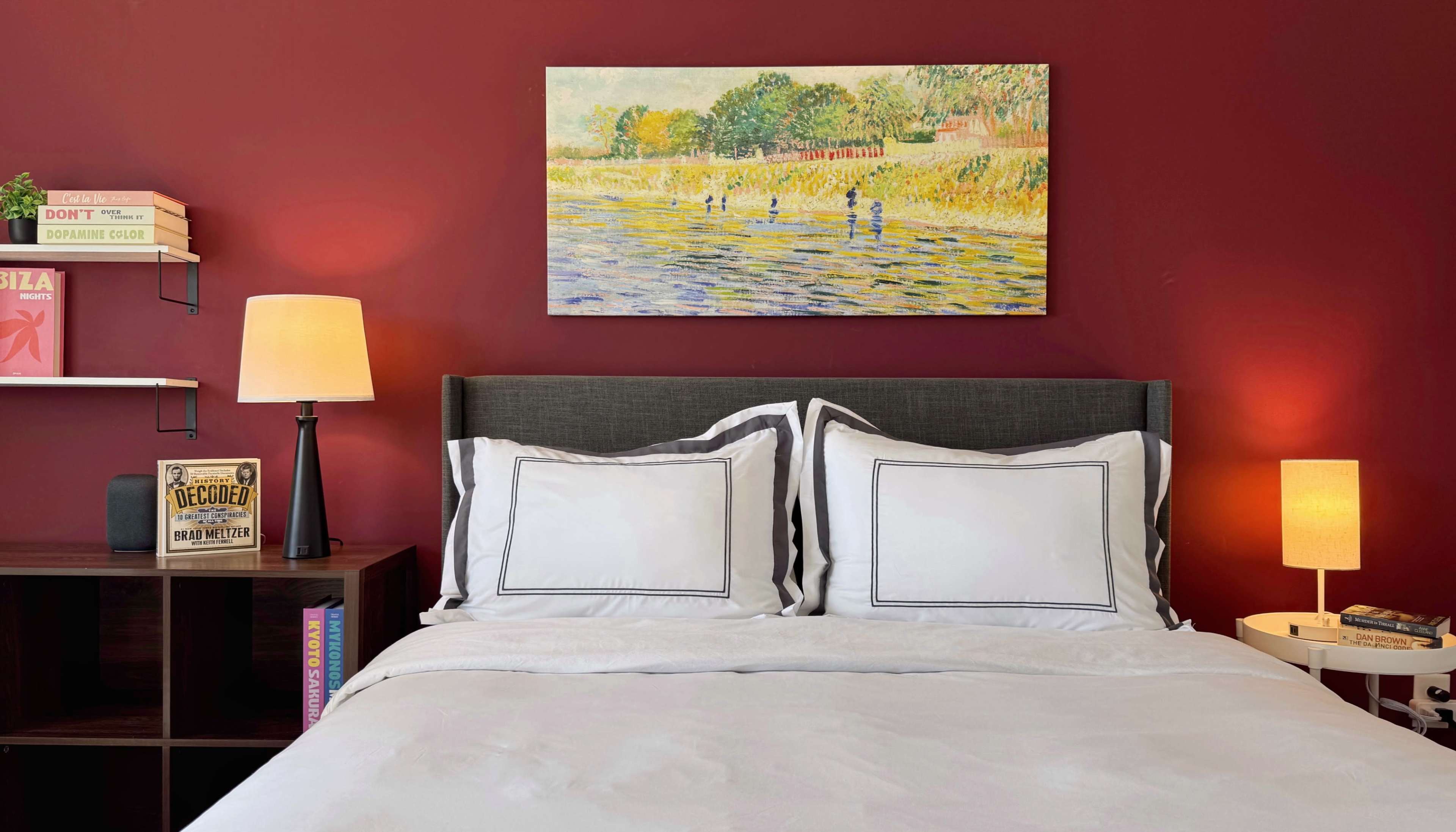 The image shows a neatly made bed with white linens and black-bordered pillows, accompanied by a bedside lamp and a painting of figures by a river on a deep red wall.