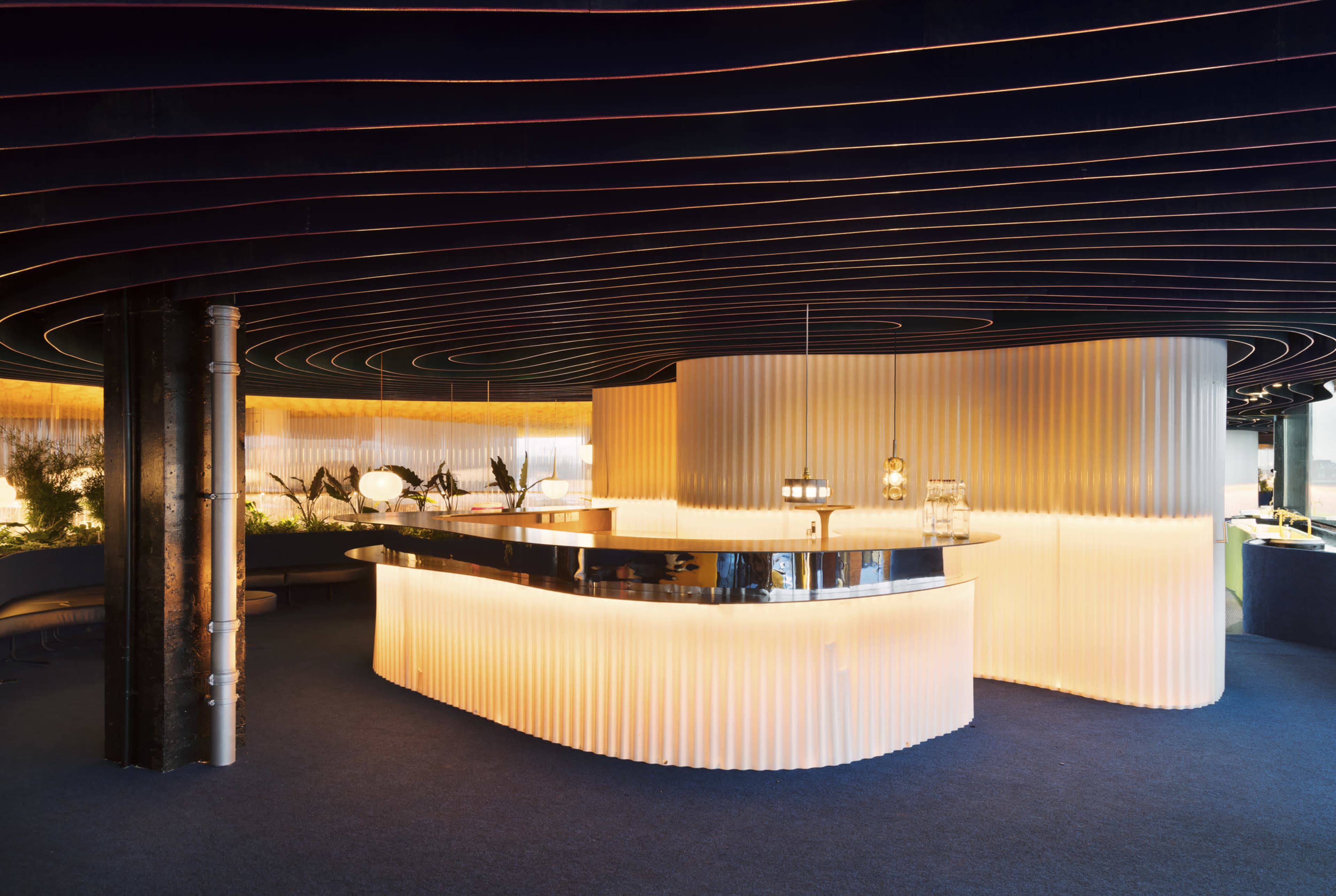 The image shows a modern bar area with a curved light-colored counter, featuring a black bar top and illuminated by warm lighting against a backdrop of dark, wavy ceiling details.