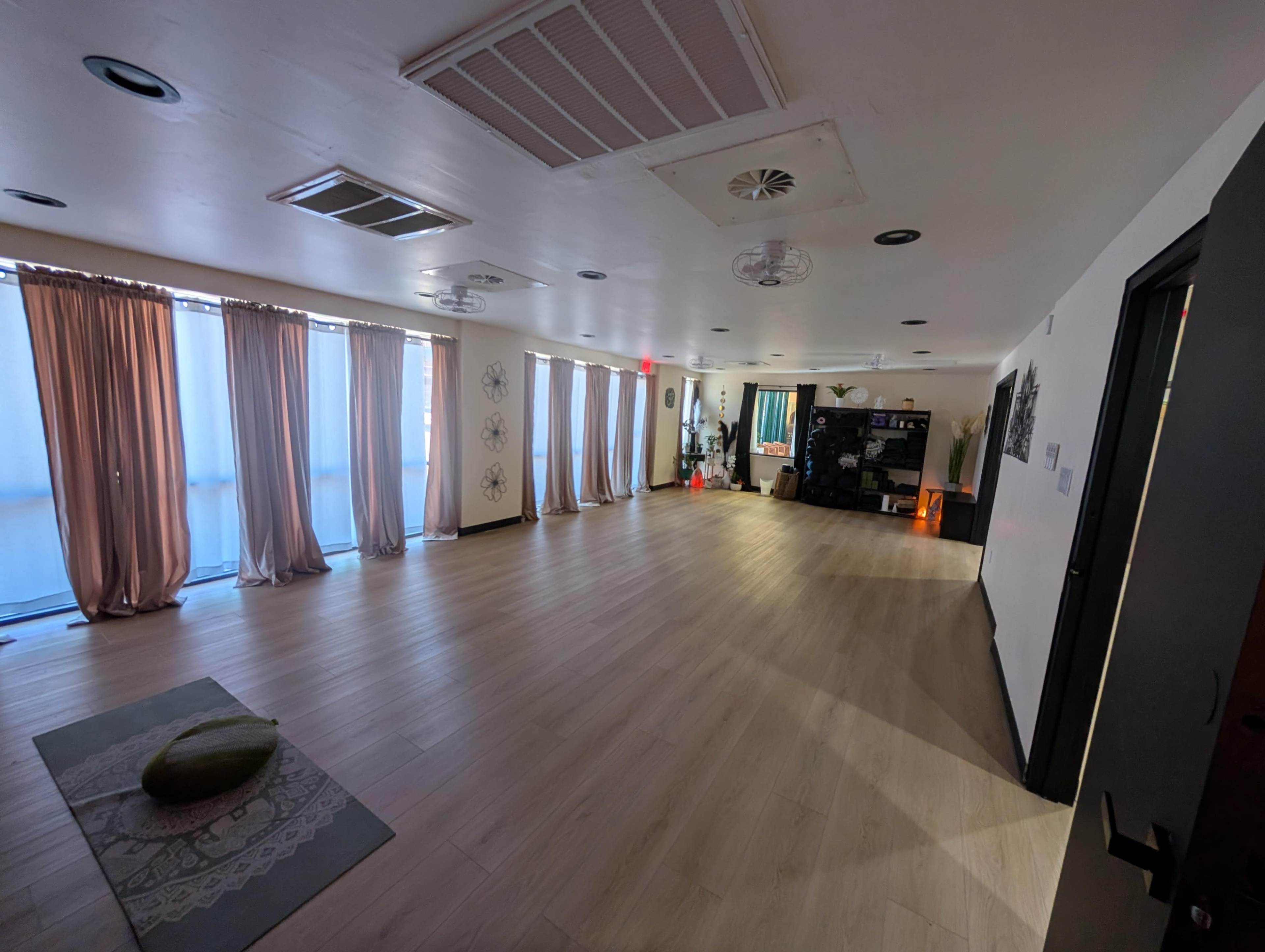 The image shows a spacious, minimally furnished room with light-colored flooring, large windows covered by pink curtains, and a few decorative elements along the walls.