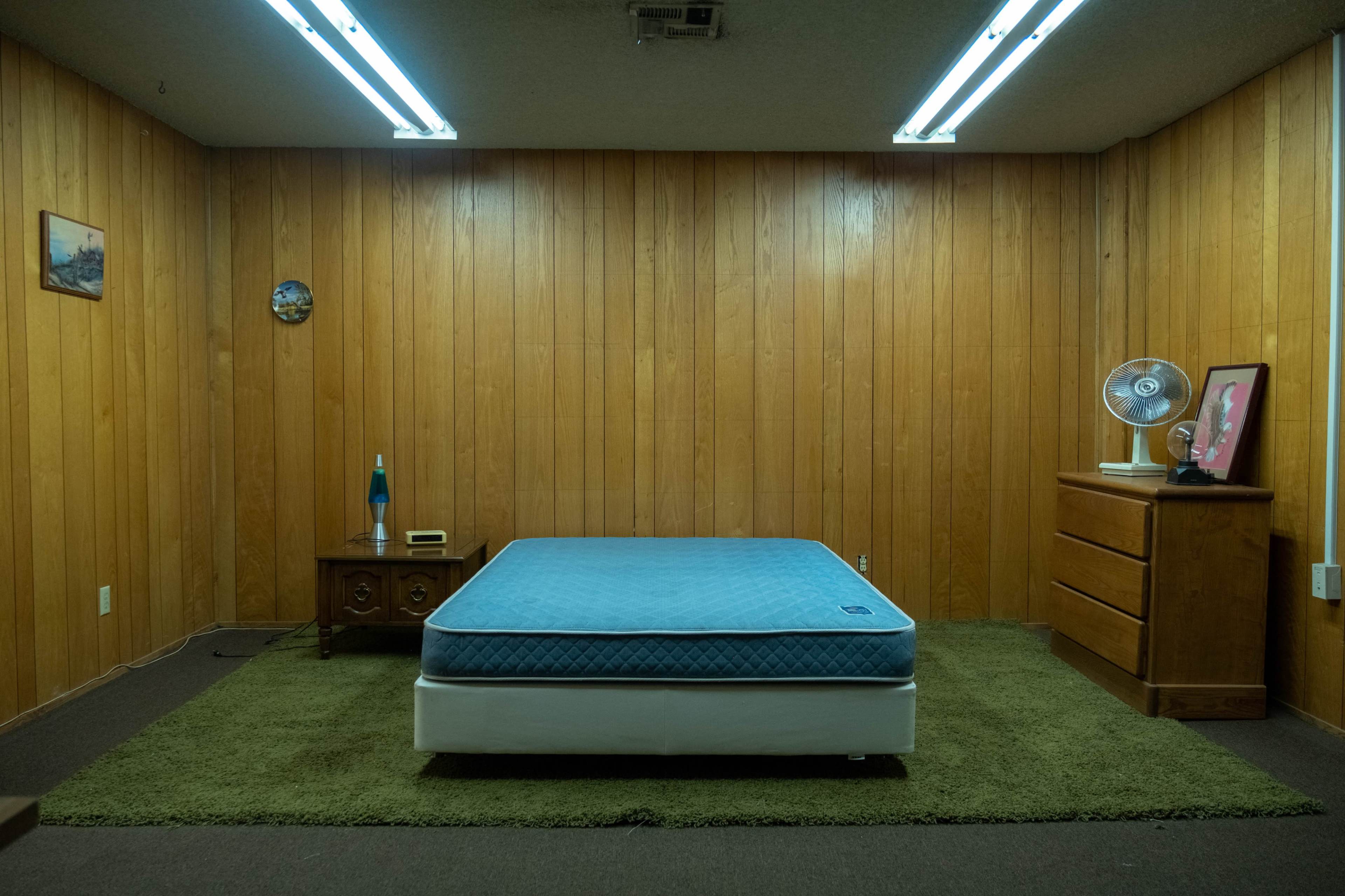 The image shows a sparsely furnished room with wood paneled walls, a blue mattress resting on a white base, a green carpet, a fan, and a dresser.