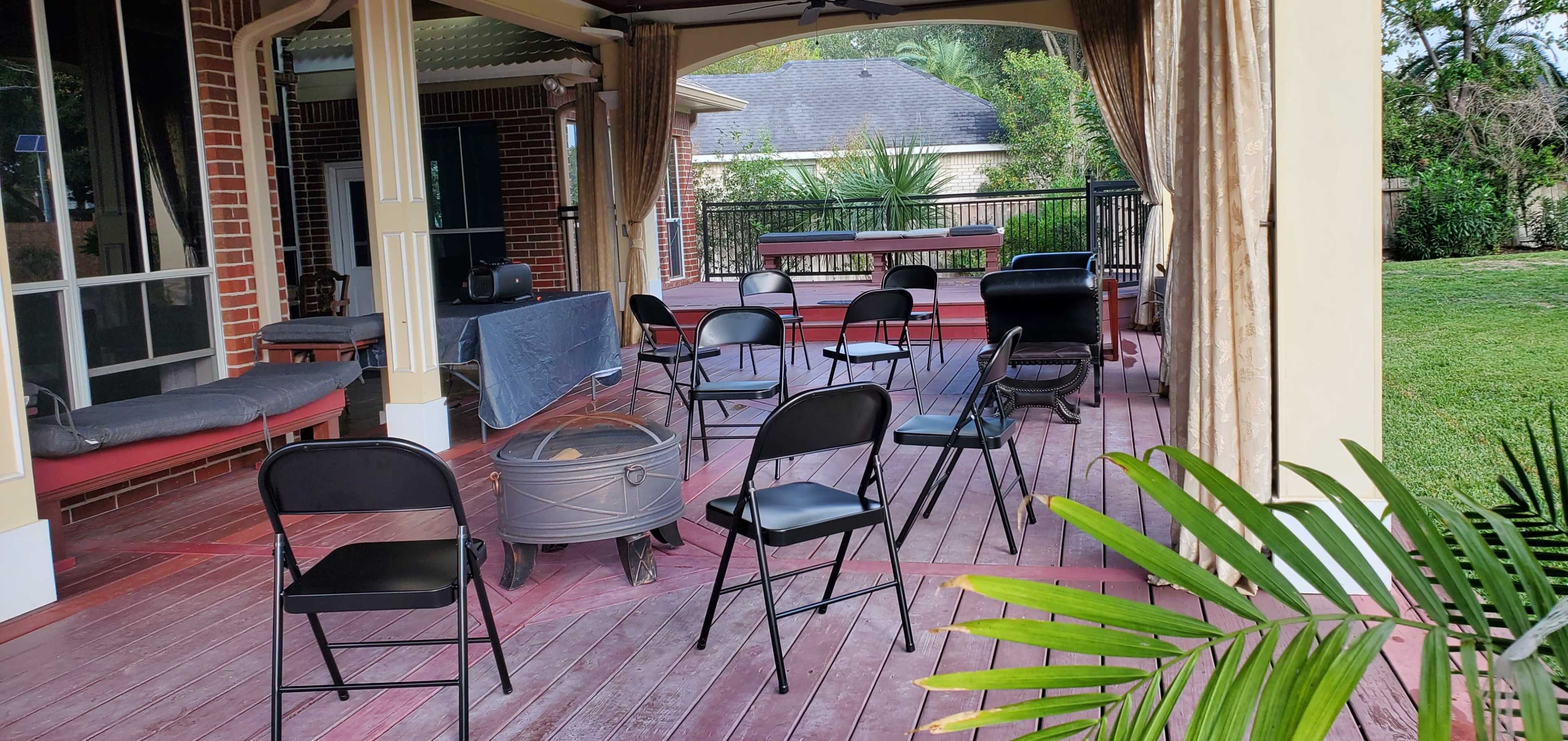 A porch area with several black folding chairs arranged around a circular fire pit on a wooden deck.