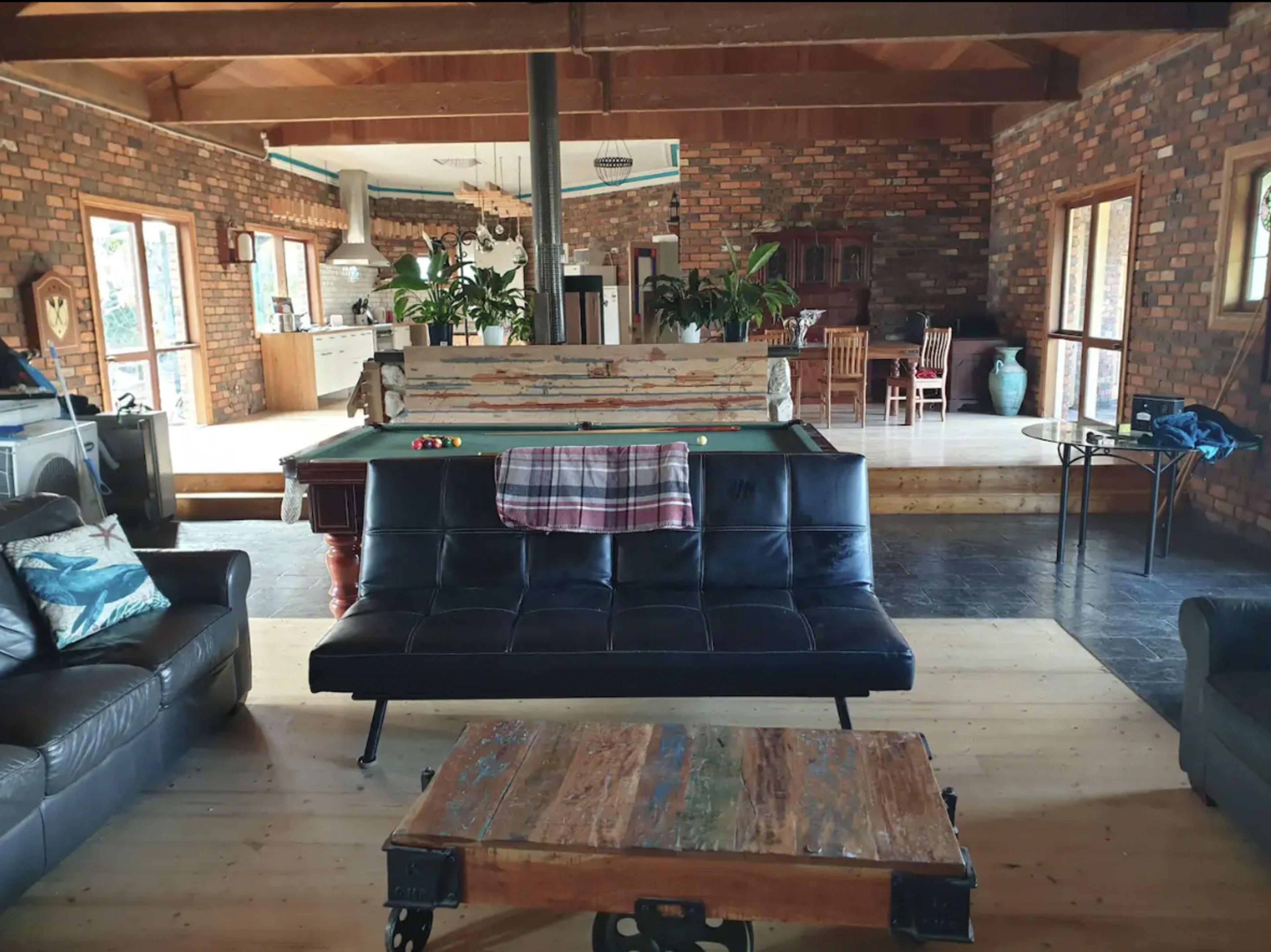A spacious interior with a pool table at the center, surrounded by a sofa and wooden furniture, against a backdrop of brick walls and large windows.