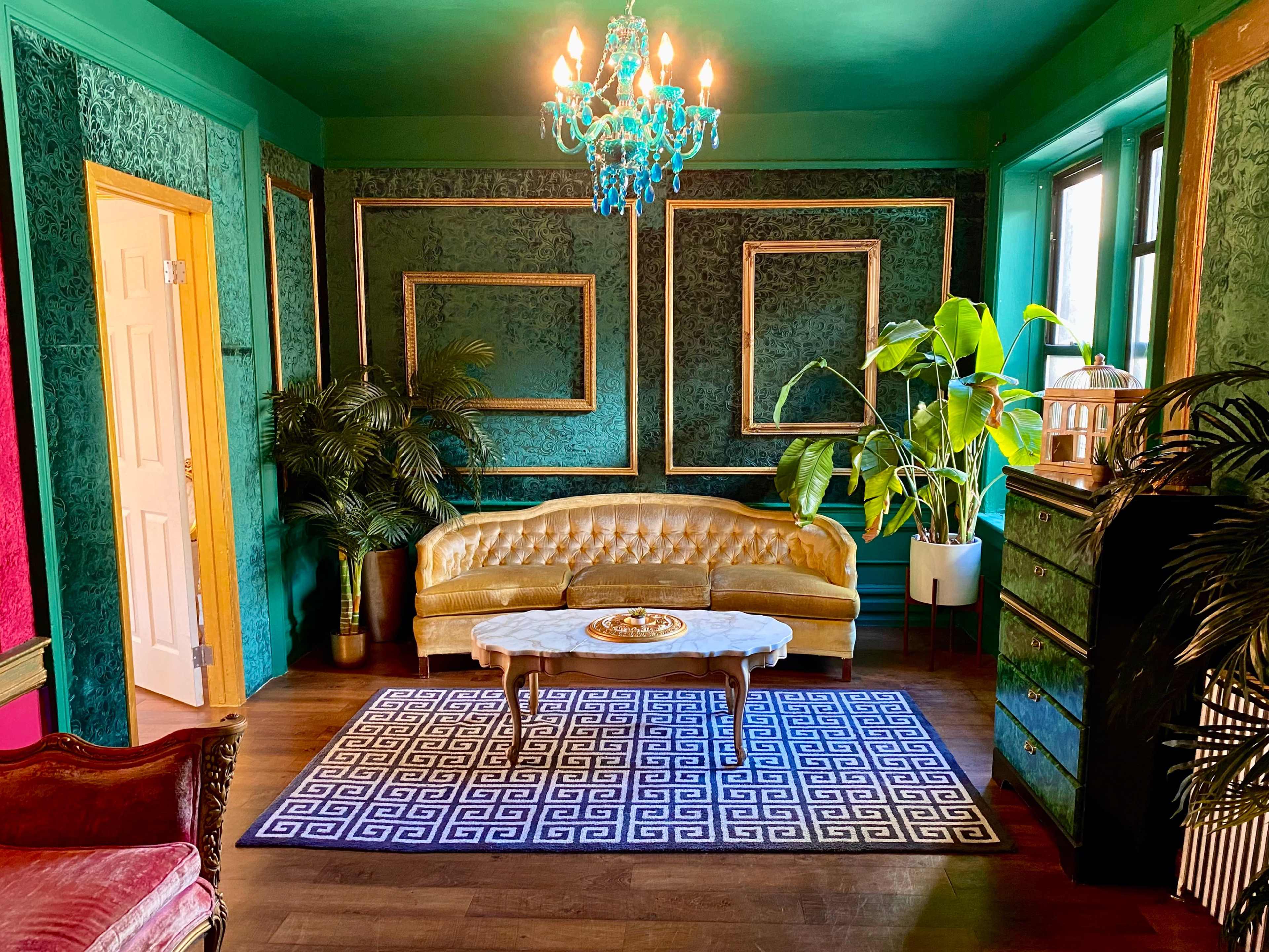 The room features a green backdrop with a gold-framed couch, a chandelier, and various plants, arranged around a coffee table on a patterned rug.