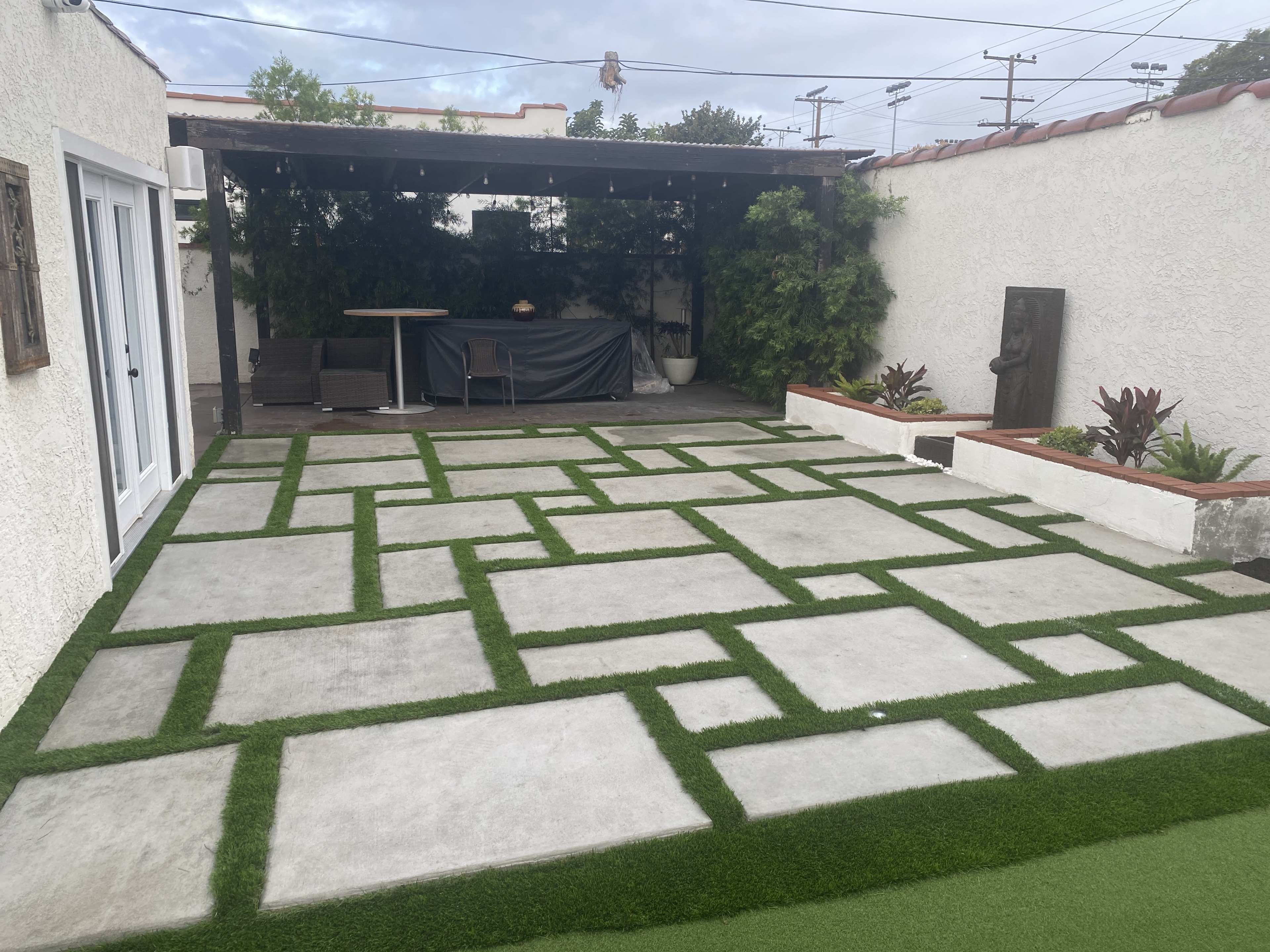 A rectangular patio with a geometric pattern of concrete slabs surrounded by artificial grass and a covered seating area in the background.