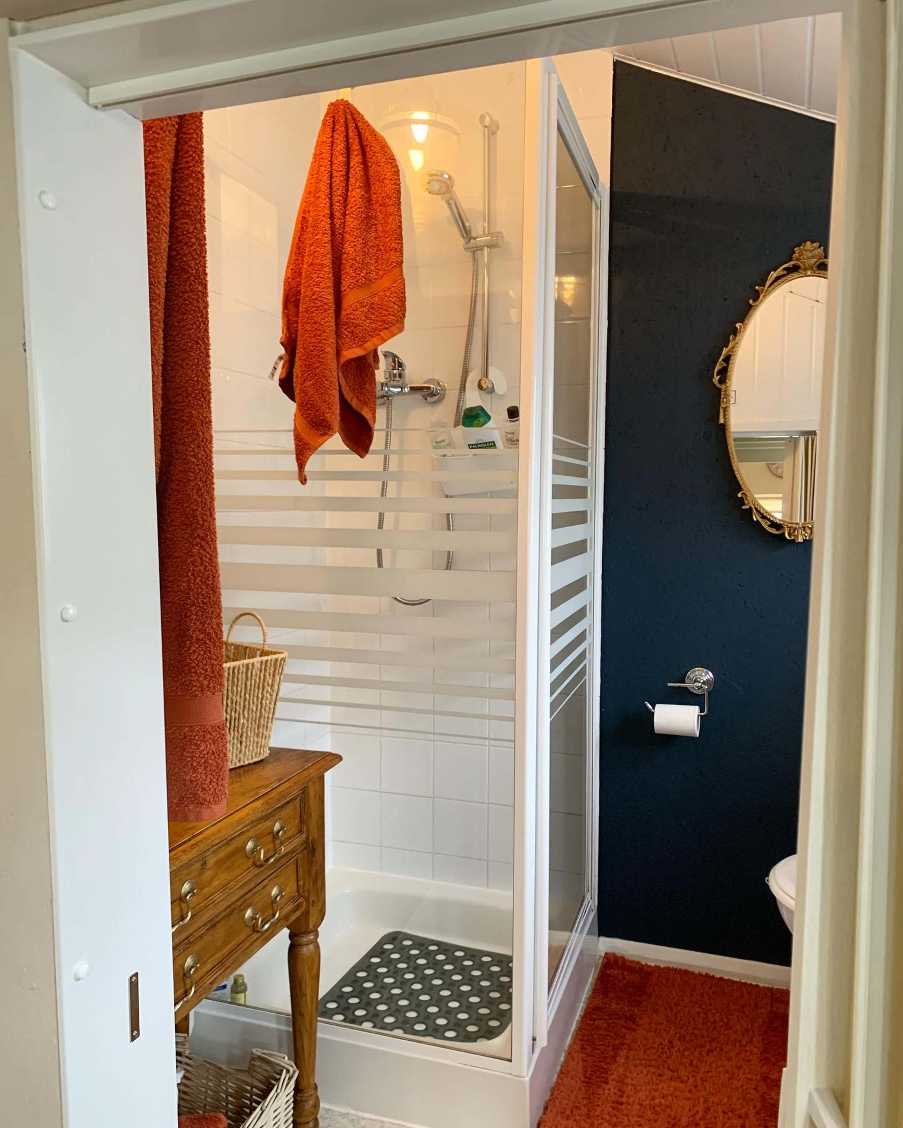 A bathroom features a clear shower stall with striped glass, an orange towel hanging nearby, a decorative mirror on the wall, and a wooden cabinet with a woven basket.