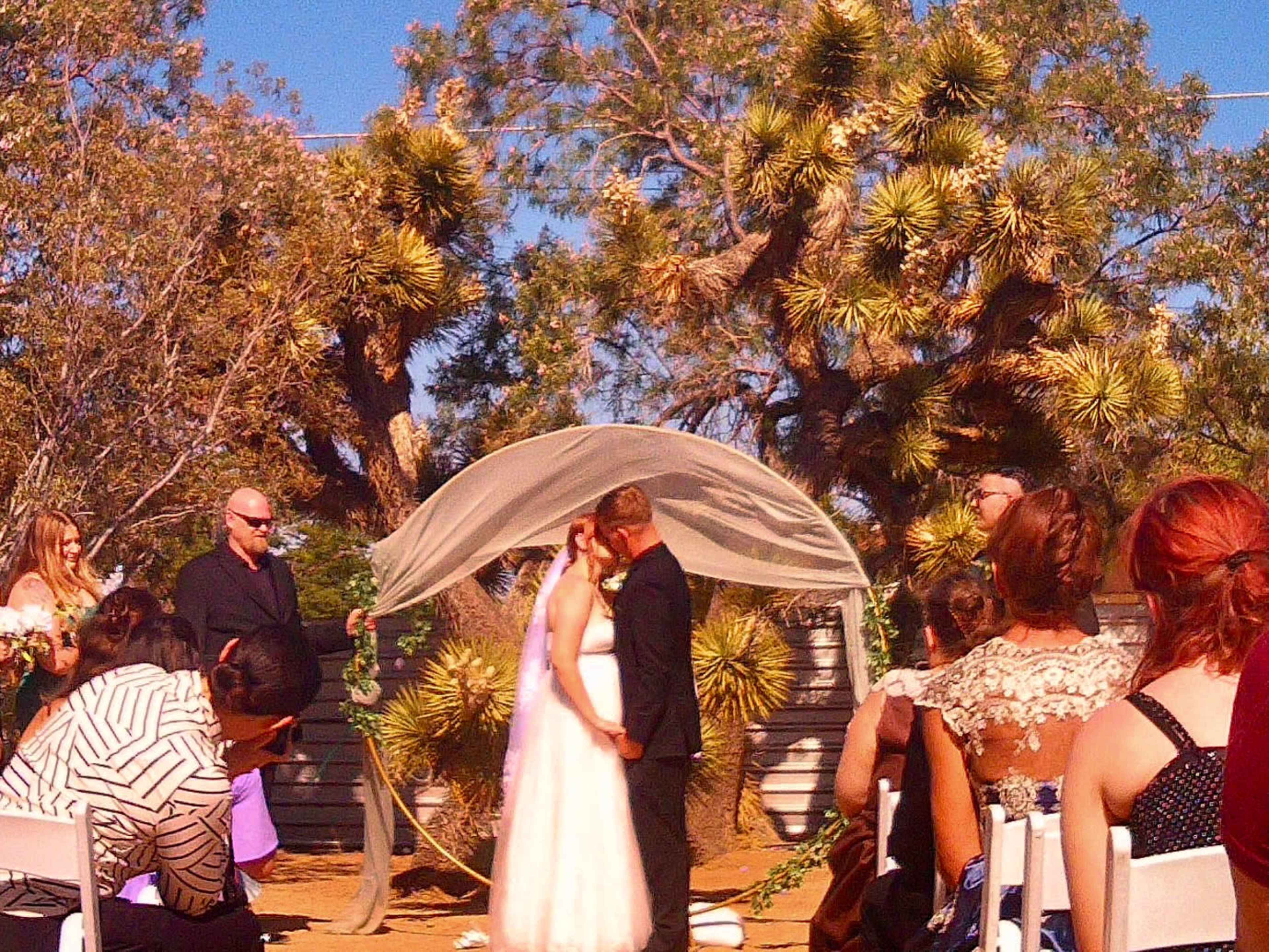 A couple shares a kiss under a canopy during their outdoor wedding ceremony with guests in the background.