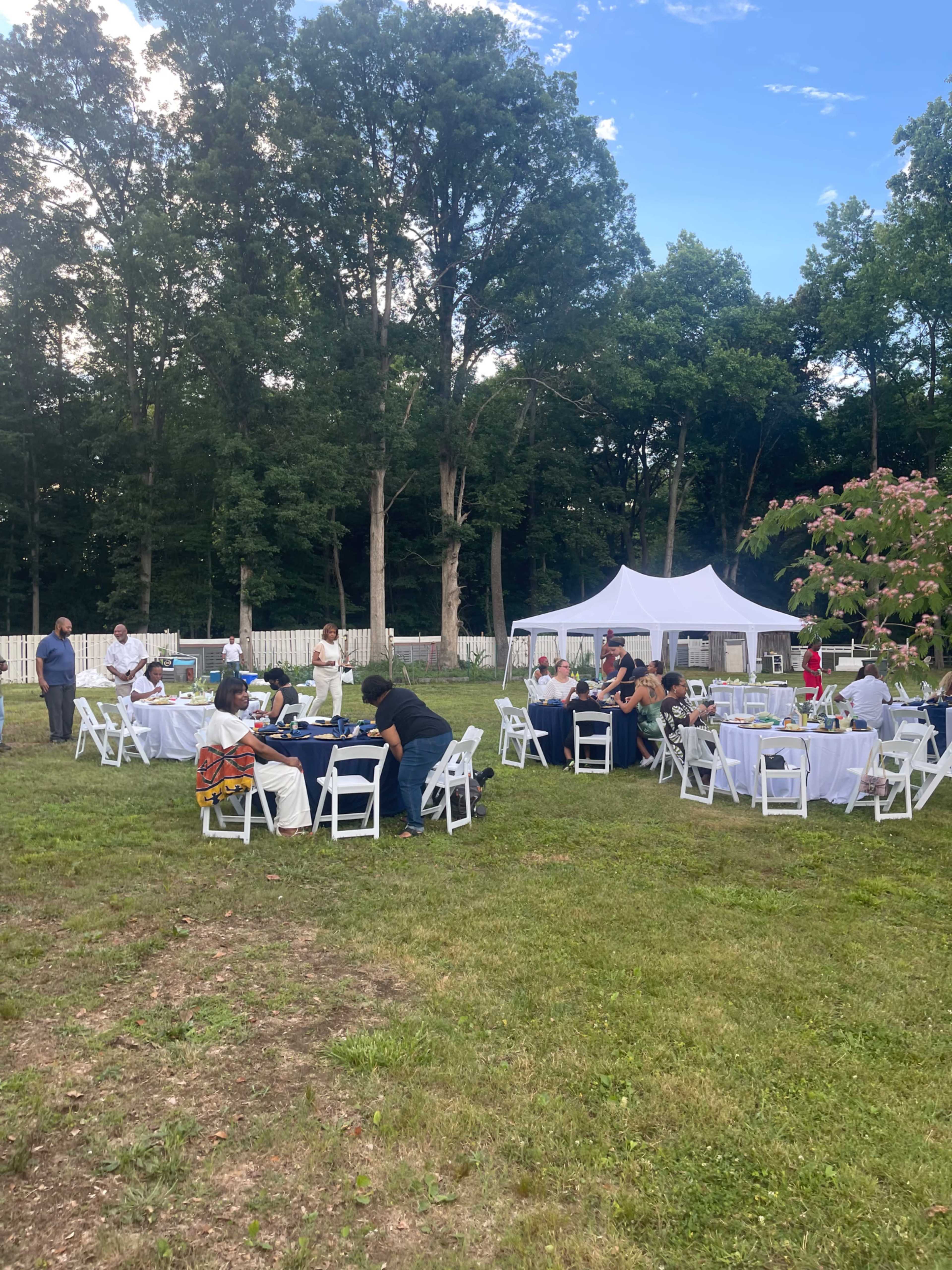A gathering of people sits at tables under a tent in a green outdoor area surrounded by trees.