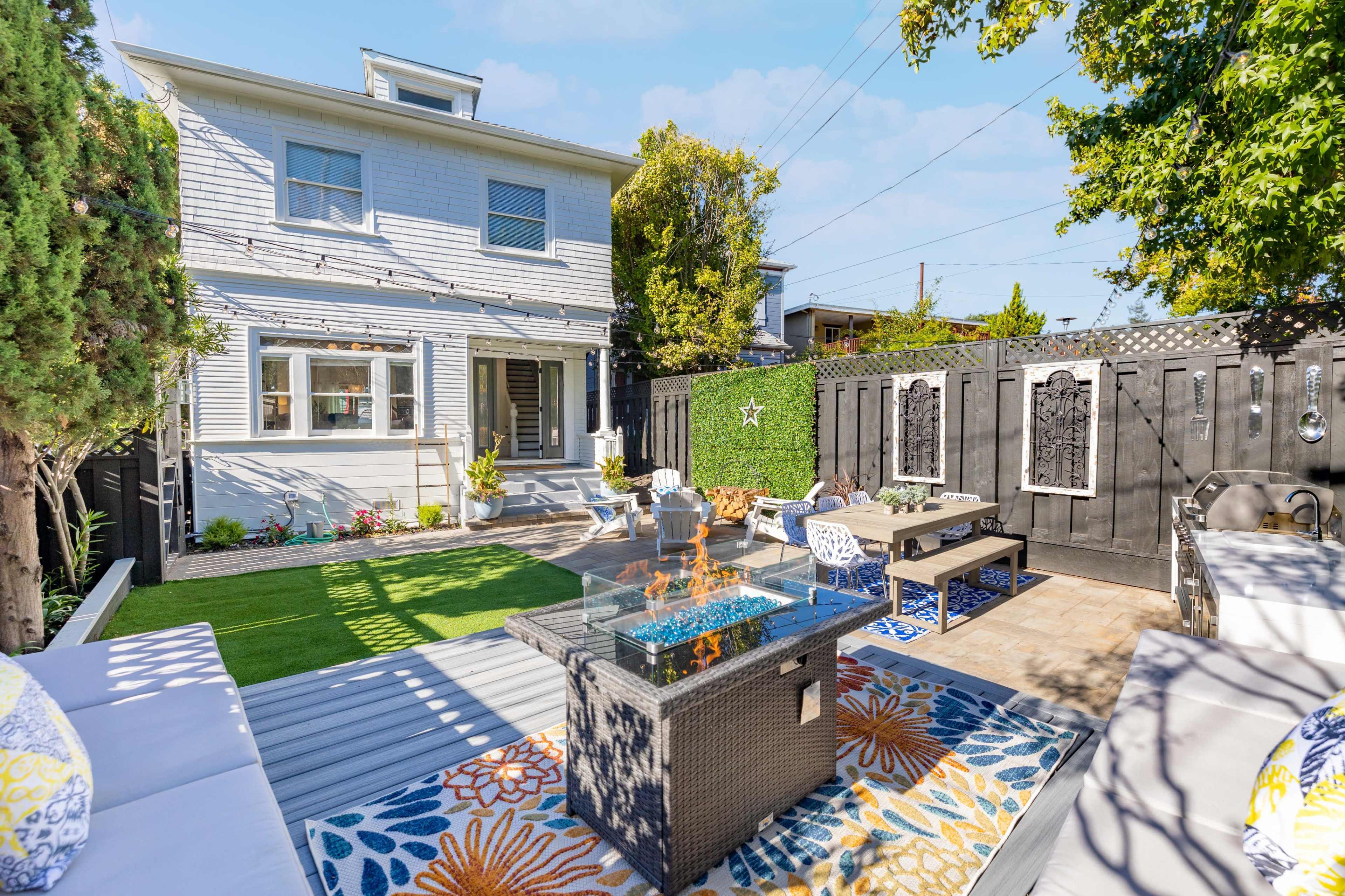 A backyard with a fire pit on a patio surrounded by seating areas, a green lawn, and a two-story house in the background.