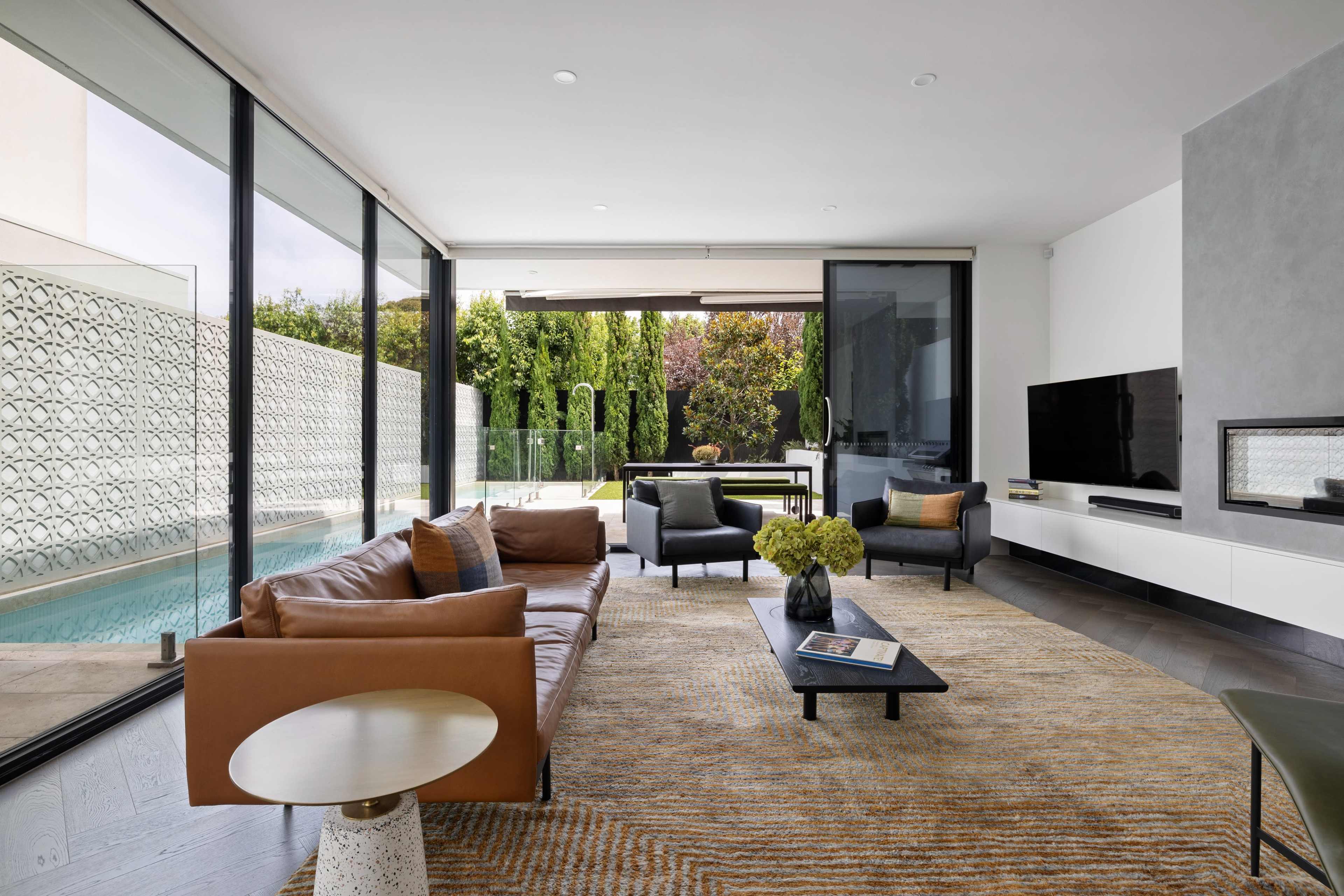 A modern living room with a brown leather sofa, black armchairs, a coffee table, and large glass windows overlooking a pool and garden.