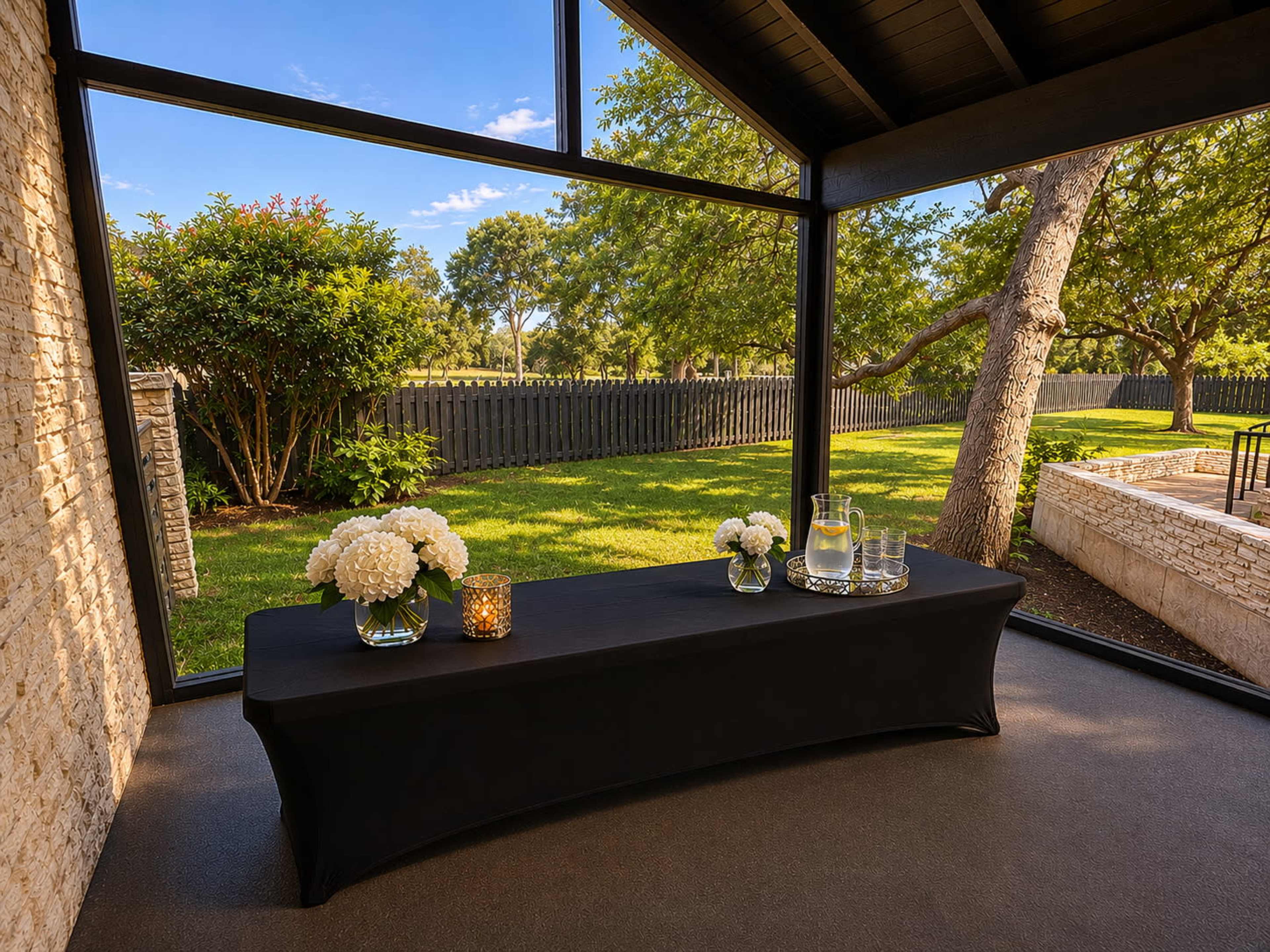 A black table draped with floral arrangements and a pitcher of water is set against a backdrop of greenery and a clear blue sky visible through large windows.