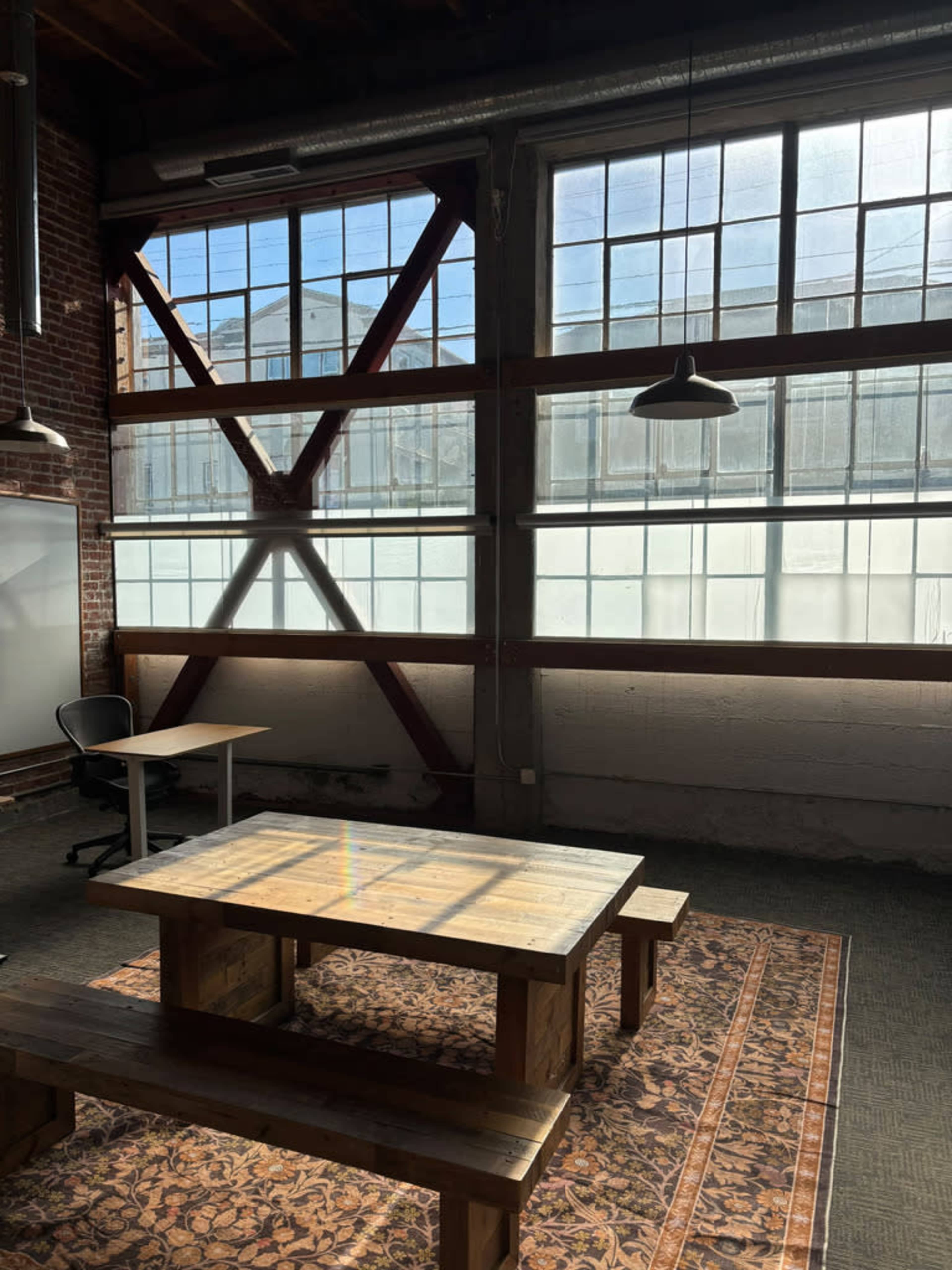 The image shows a spacious office interior with large windows, brick walls, a wooden table, and a single chair in a well-lit environment.