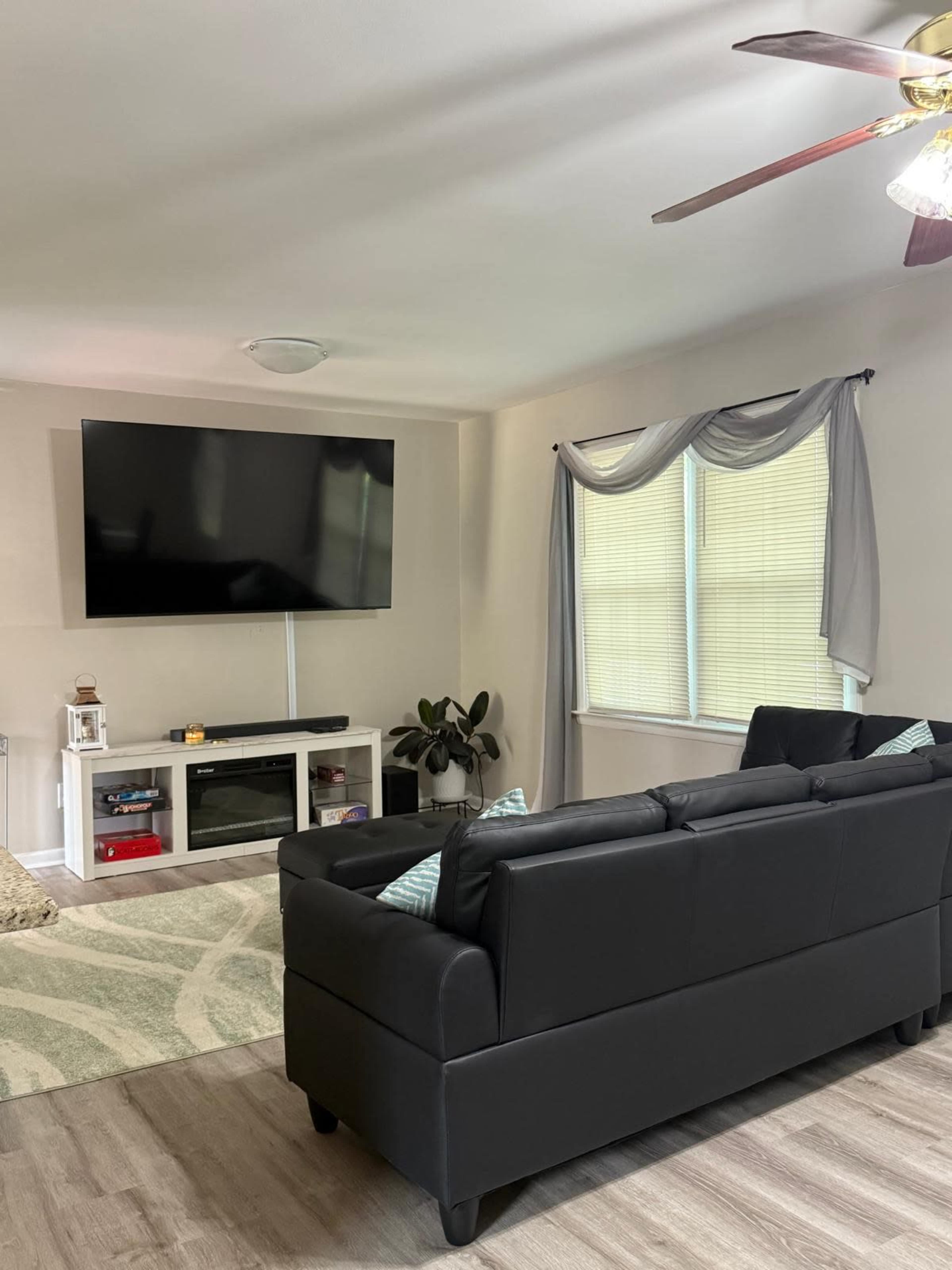 The image shows a living room with a black sofa facing a large wall-mounted television, a white entertainment unit, and a window with sheer curtains.