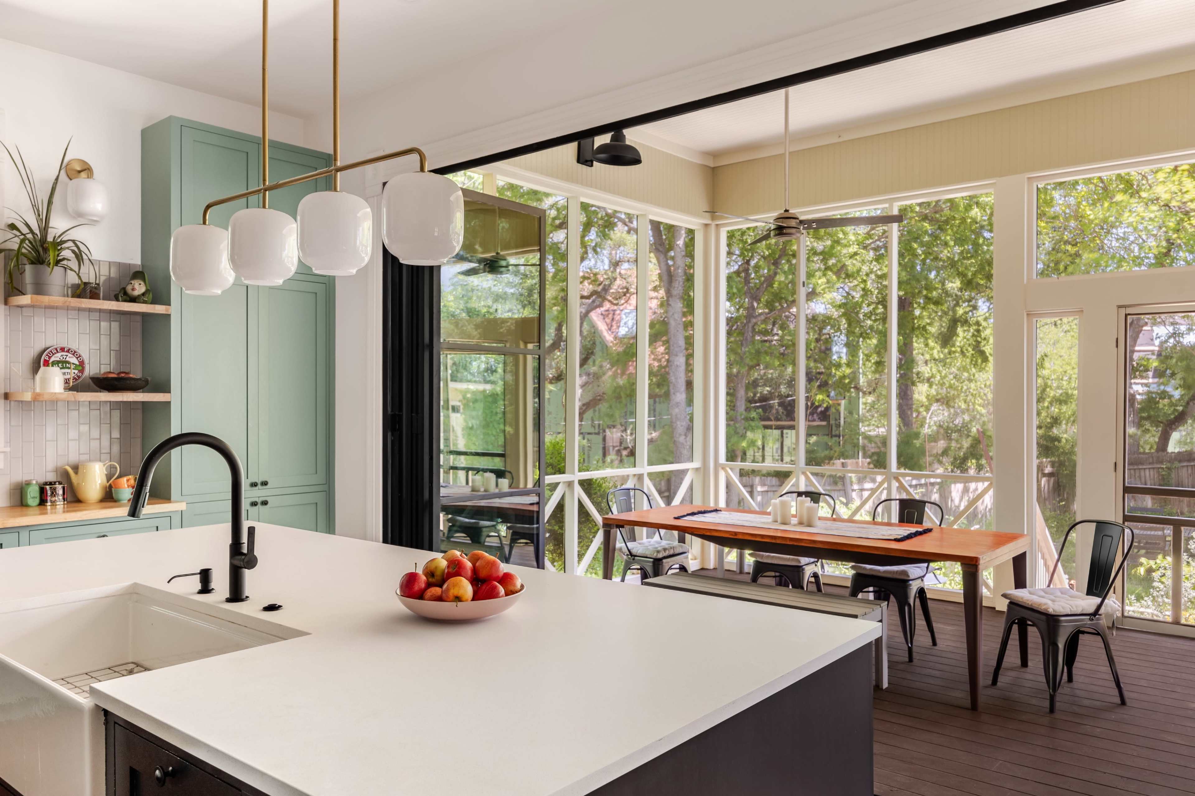 The image shows a modern kitchen with a white island, a bowl of apples, and large windows that open to a screened porch area with a dining table.