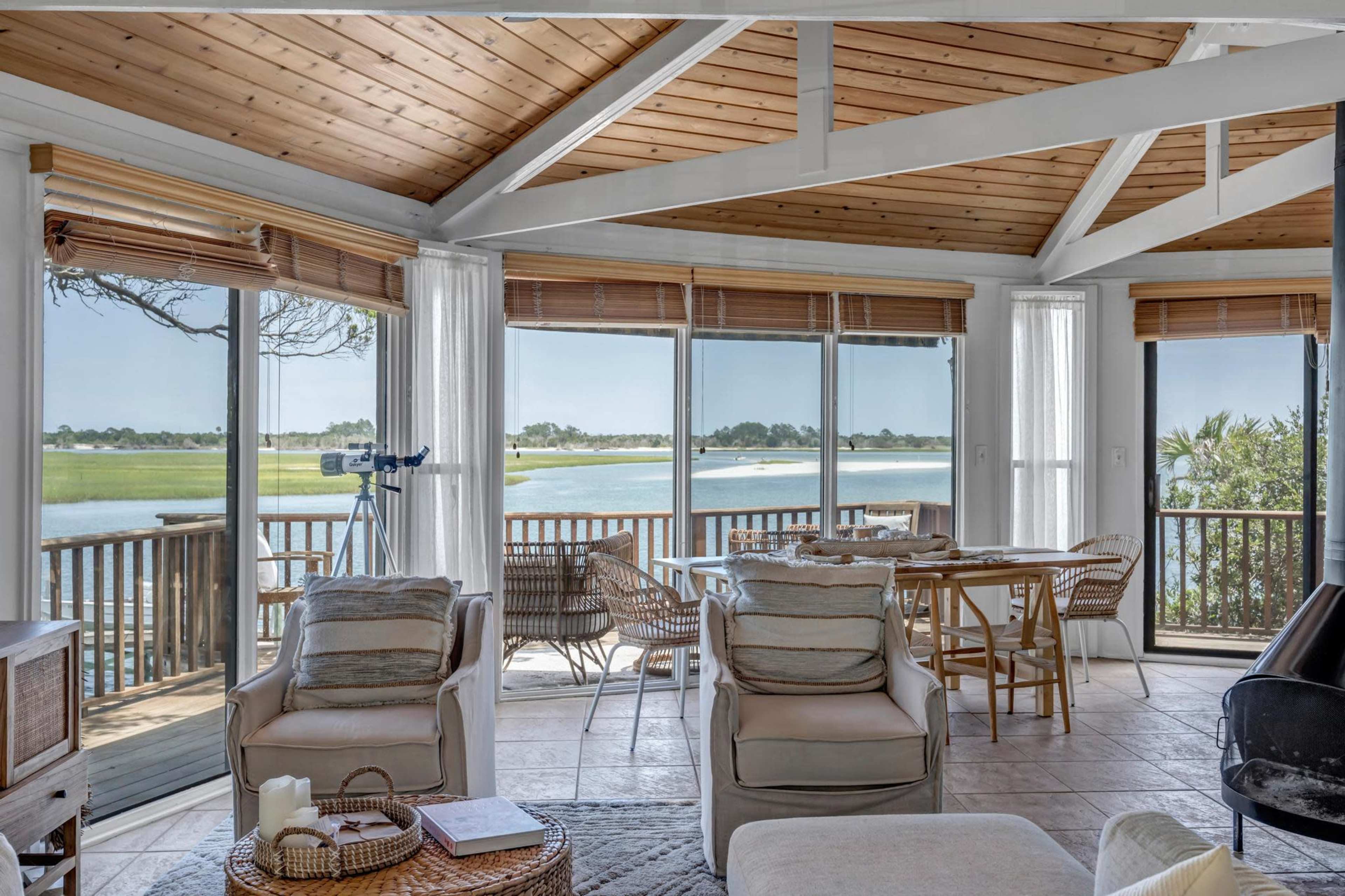 A sunlit interior of a living space with large windows overlooking a serene body of water, featuring comfortable seating and a dining area.