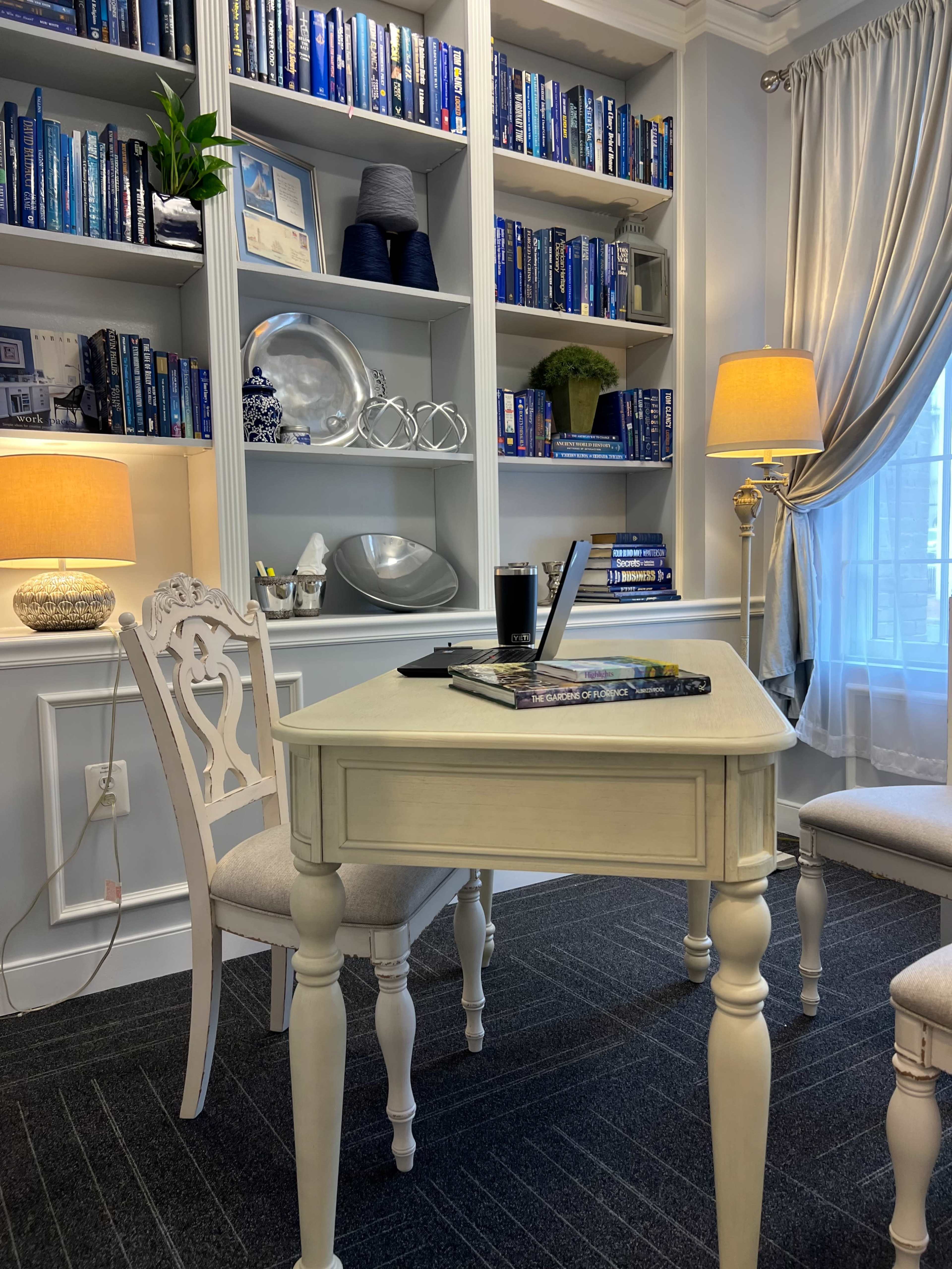 A small white table with a book and a drink sits in front of a bookshelf filled with blue-covered books, accompanied by a chair and two lamps illuminating the room.