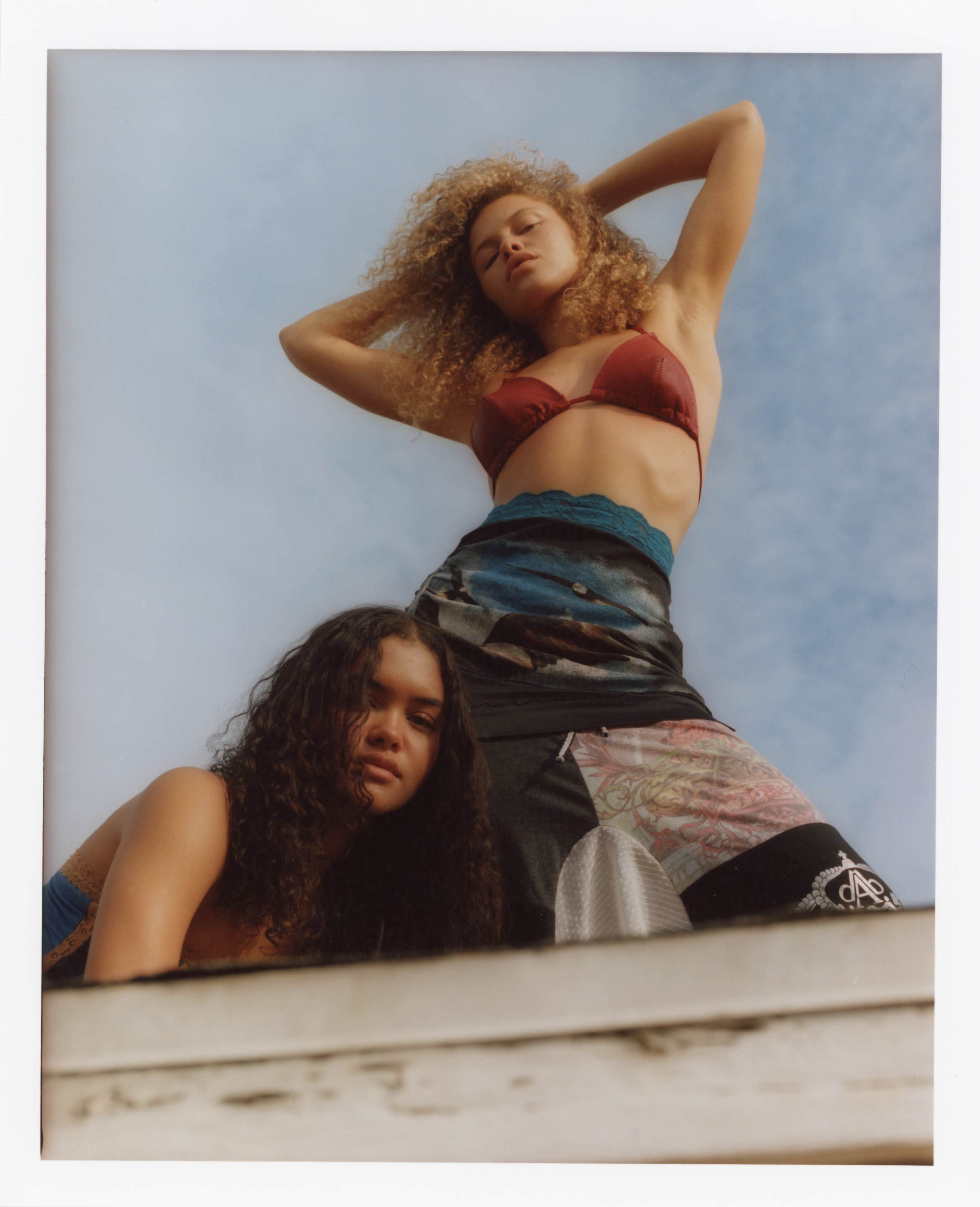 Two young women pose outdoors, one standing above the other while showcasing summer fashion against a blue sky.