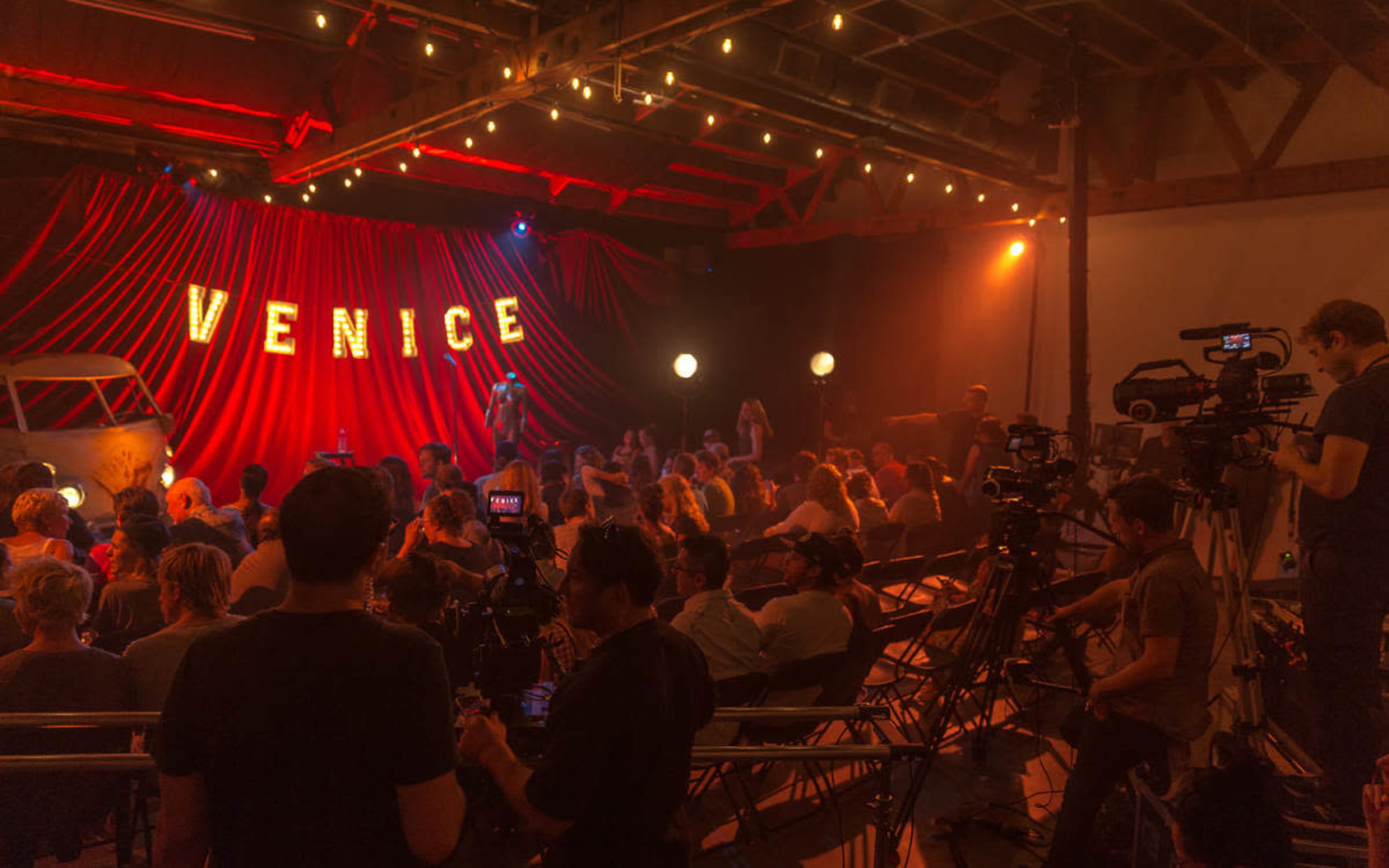 A crowd sits in a dimly lit venue with a stage featuring the word "VENICE" illuminated in bright letters.