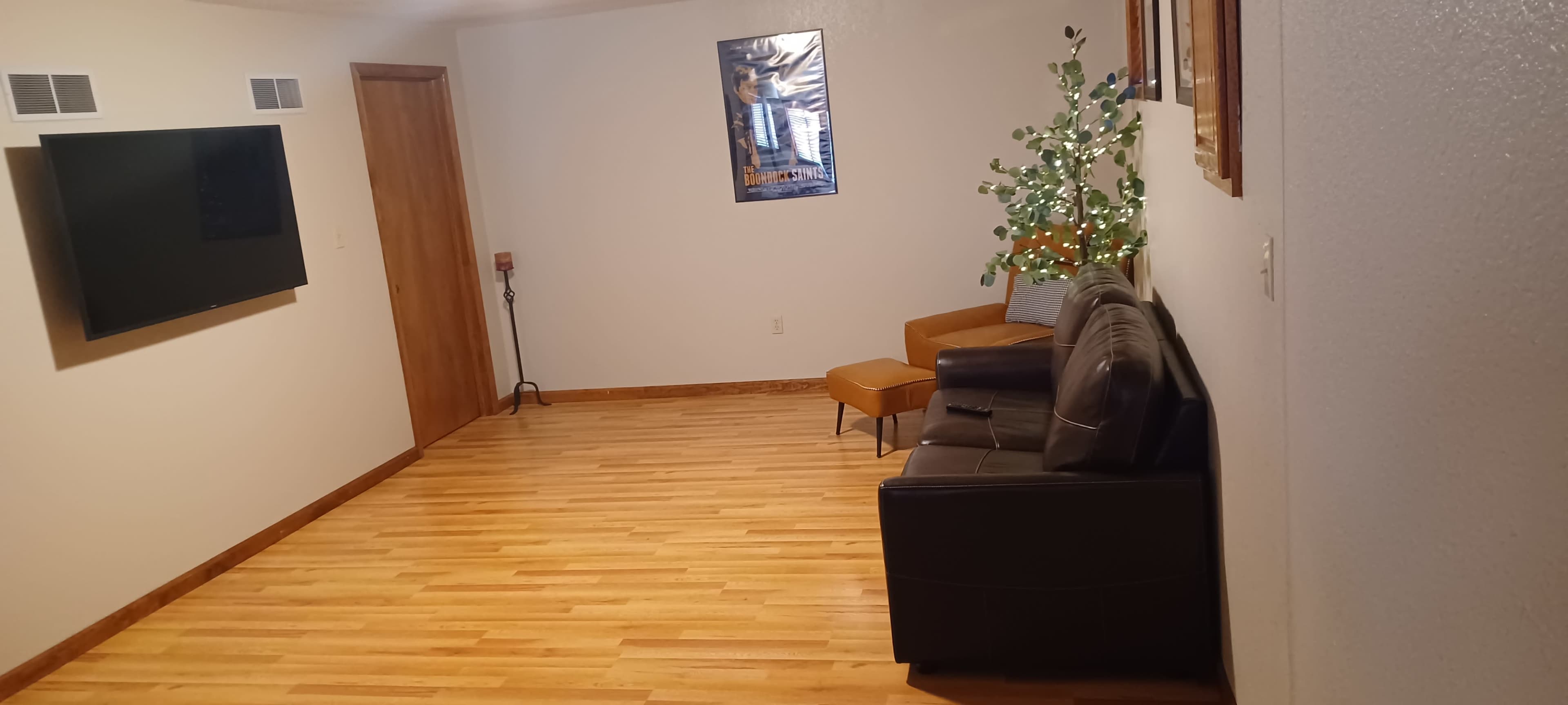 The image shows a living room with a television on the wall, a brown sofa, an orange accent chair, and a decorative plant with lights.