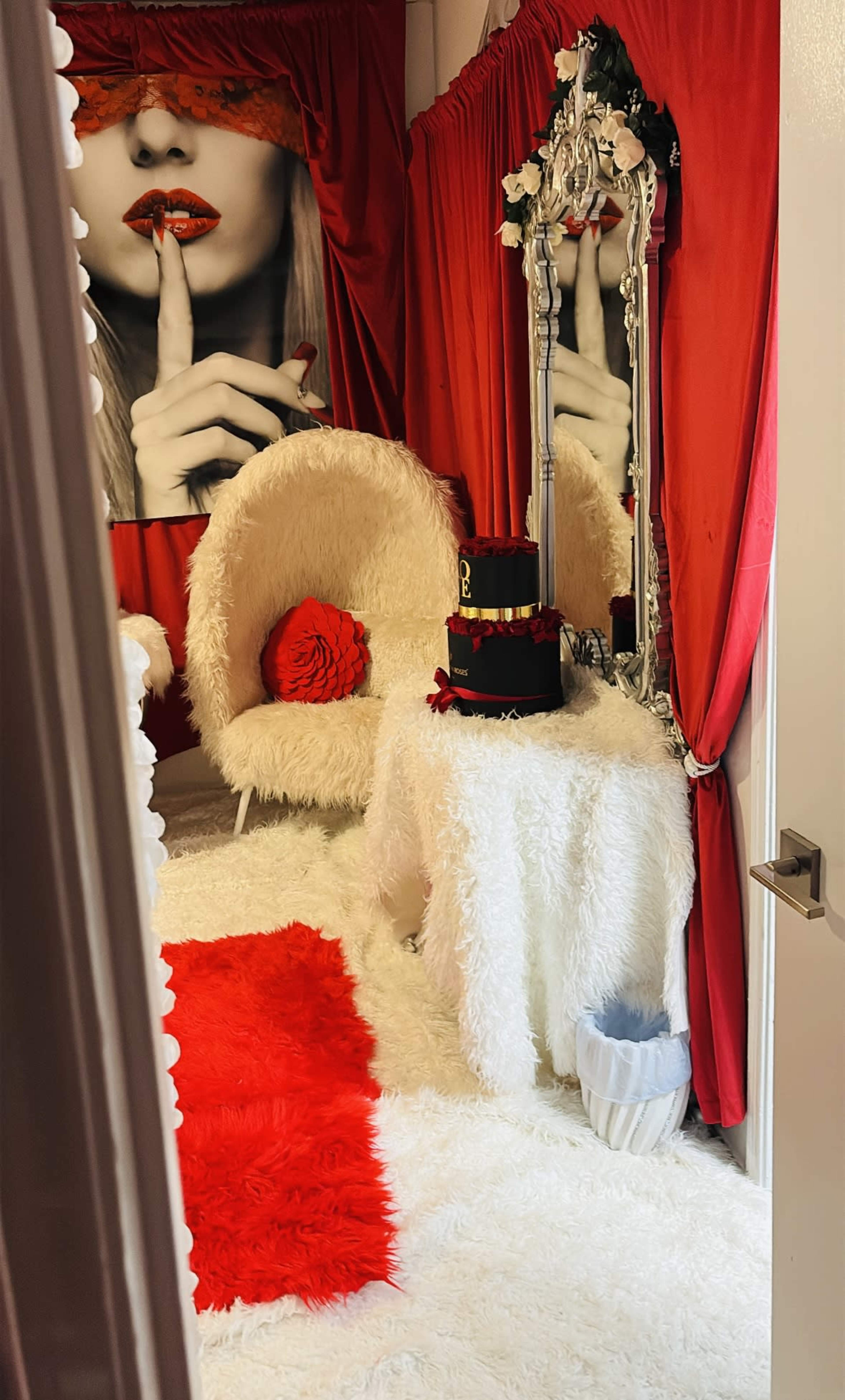 The image shows a stylish room decorated with red and white furnishings, including a fluffy white chair, a round table, and a mirror, all set against a backdrop of red curtains and a large black-and-white wall print.