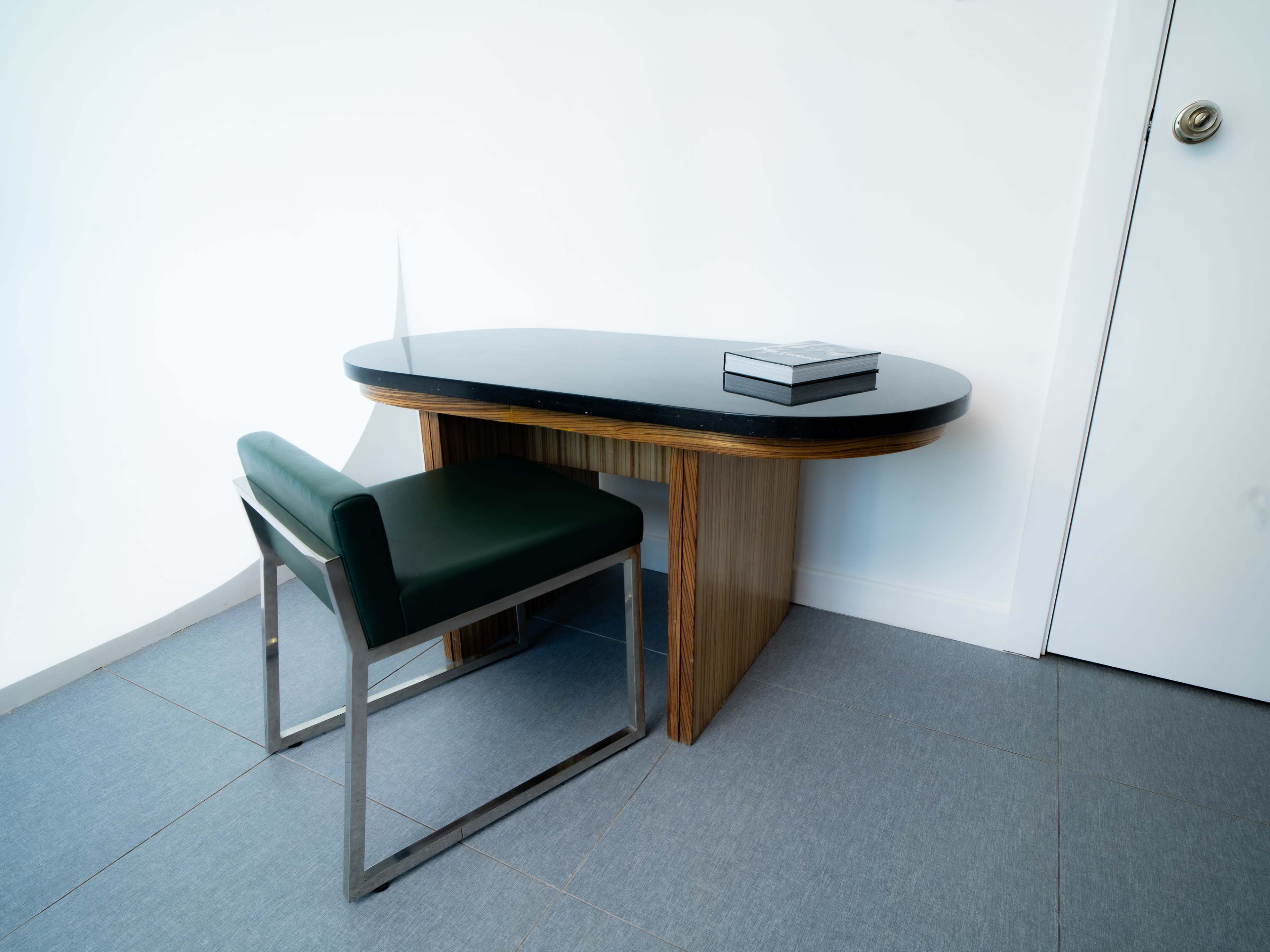 A sleek, modern desk with a black top is positioned next to a green chair, set against a minimalist white wall and gray floor tiles.