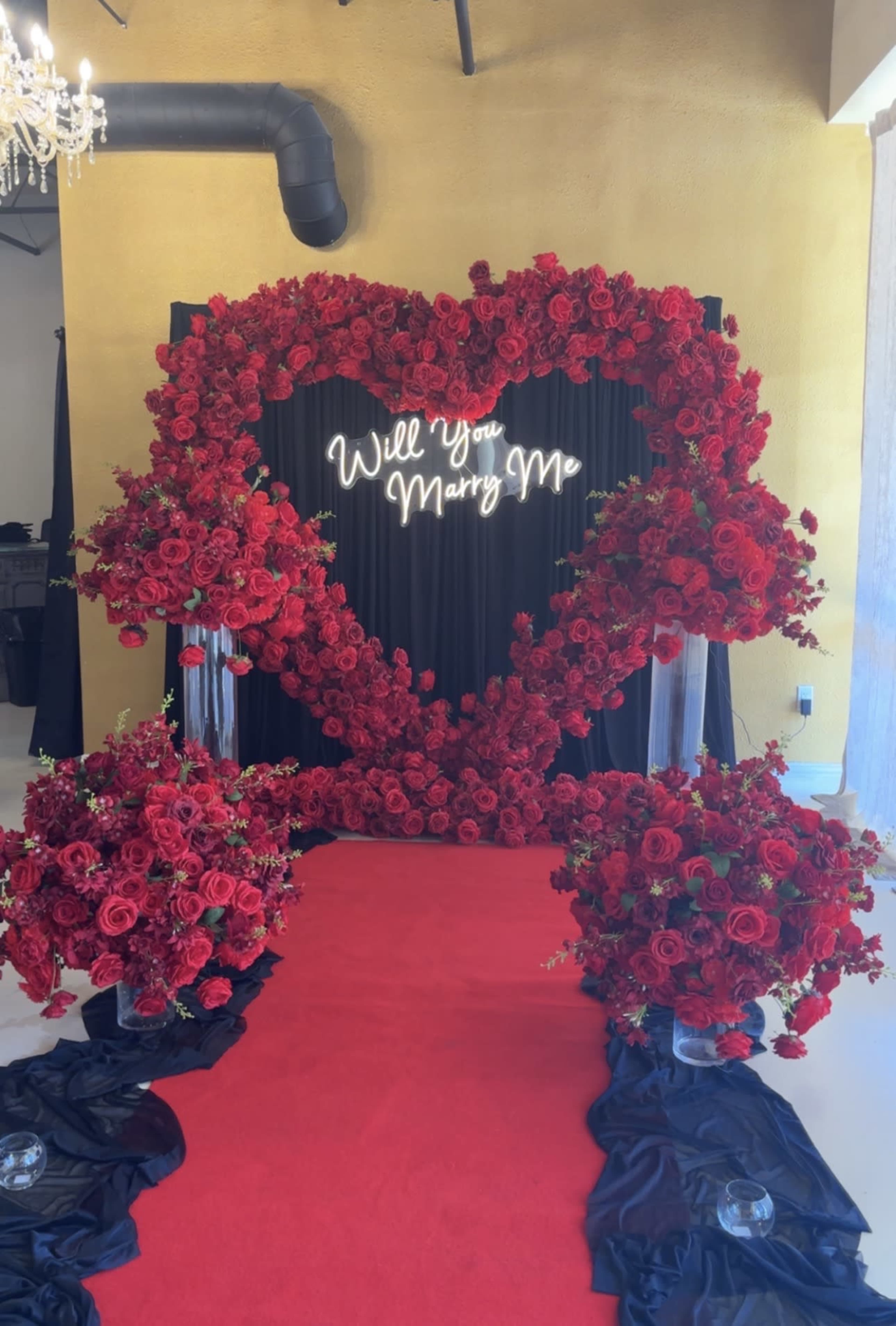 A heart-shaped floral arrangement made of red roses frames a neon sign that says "Will You Marry Me," with additional floral displays on either side and a red carpet leading to it.
